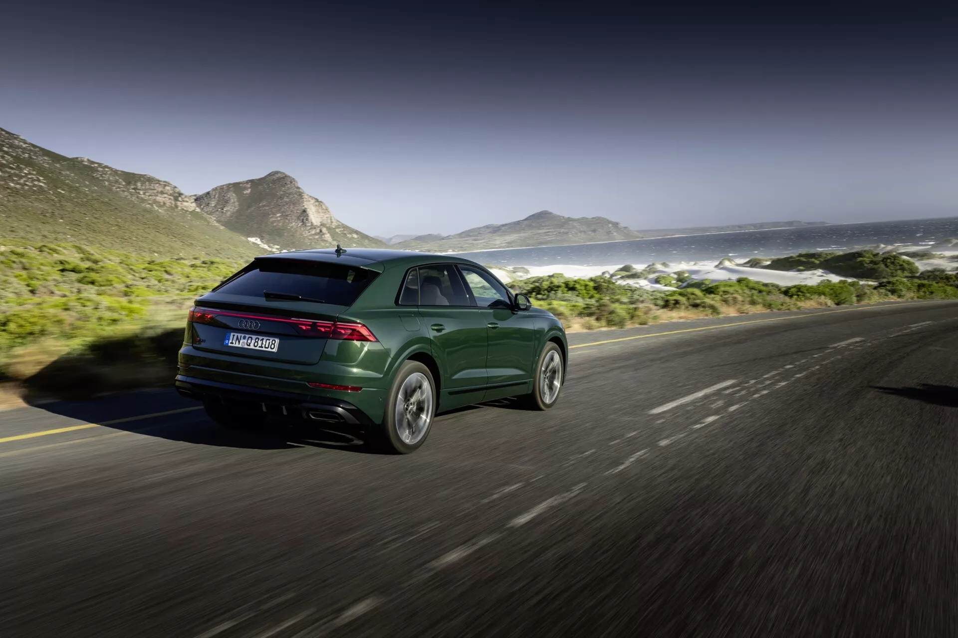 Audi Q8 2023 (4M-Facelift) v metalik temno zeleni, prikazuje svoj športen in sodoben dizajn s tankimi zadnjimi lučmi in litimi kolesi, posneto iz zadnjega tričetrtinskega kota na obalni cesti z gorami in oceansko sceno v ozadju