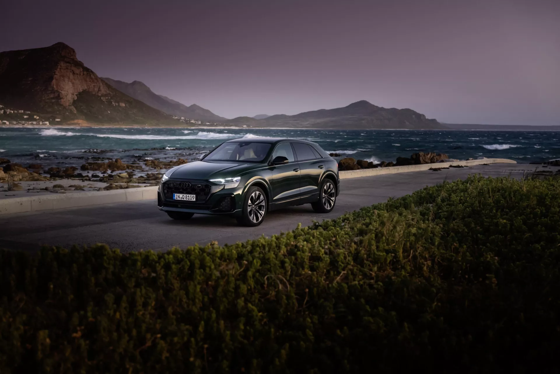 Audi Q8 2023 (4M-Facelift) temen hečbek na slikoviti obalni cesti ob mraku, ki prikazuje njegove LED žaromete, aerodinamičen profil in lita kolesa