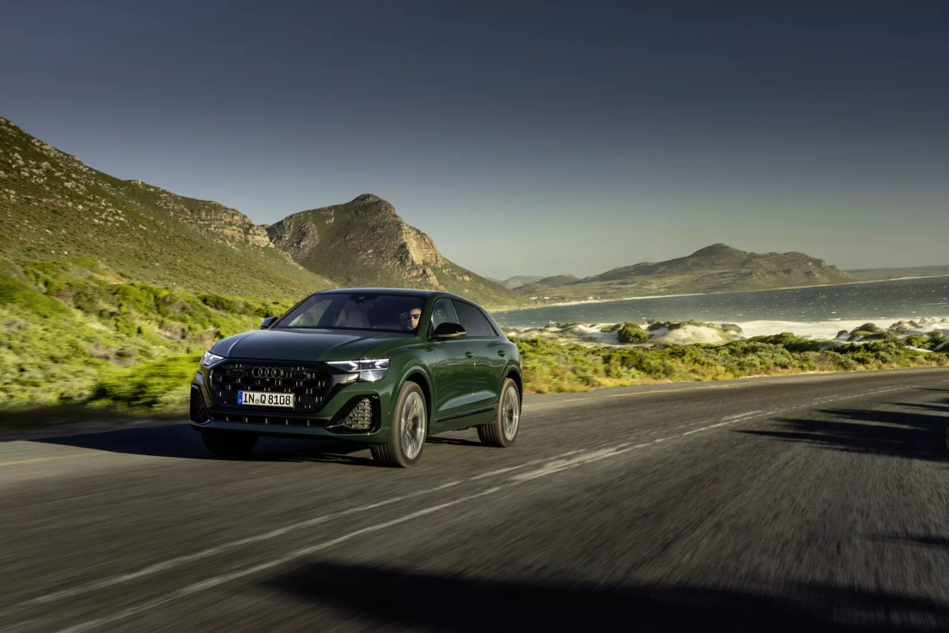 Audi Q8 4M Facelift 2023 vozi po obalni cesti z dinamičnim sprednjim pogledom, ki prikazuje LED žaromete, sodobno masko in učinek zamegljenega gibanja proti ozadju oceana in gora v sončnem dnevu