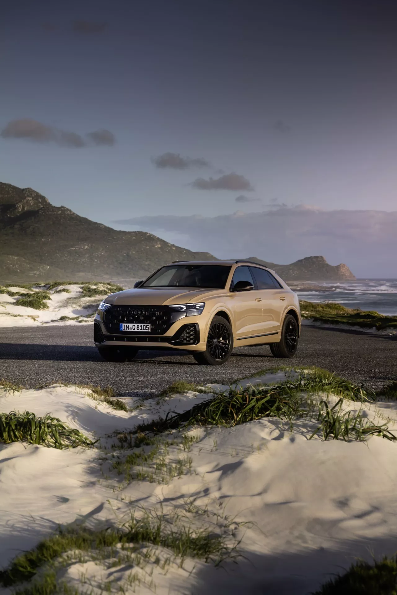 Audi Q8 2023 (4M-Facelift) v mogočni plaži in gorski pokrajini ob mraku, ki predstavlja svojo elegantno obliko in napredne funkcije, kot so LED žarometi in velika lita kolesa, iz treh četrtin sprednjega in stranskega kota