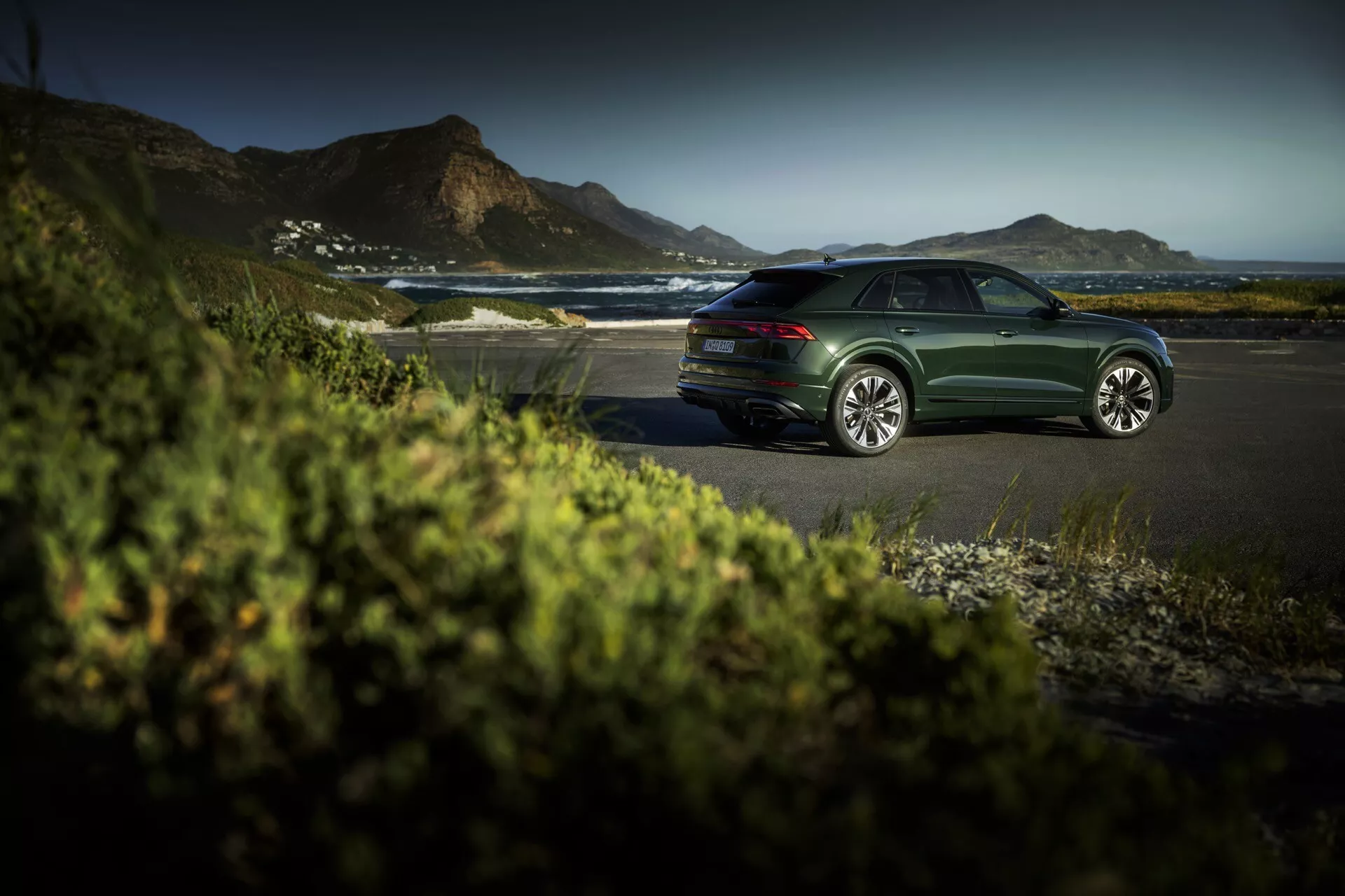 Sedan Audi Q8 (4M-Facelift) temno zelene barve parkiran na obalni cesti med zlato uro, prikazuje elegantne oblikovne linije, večkraka lita kolesa in miren luksuzni zunanji ambient