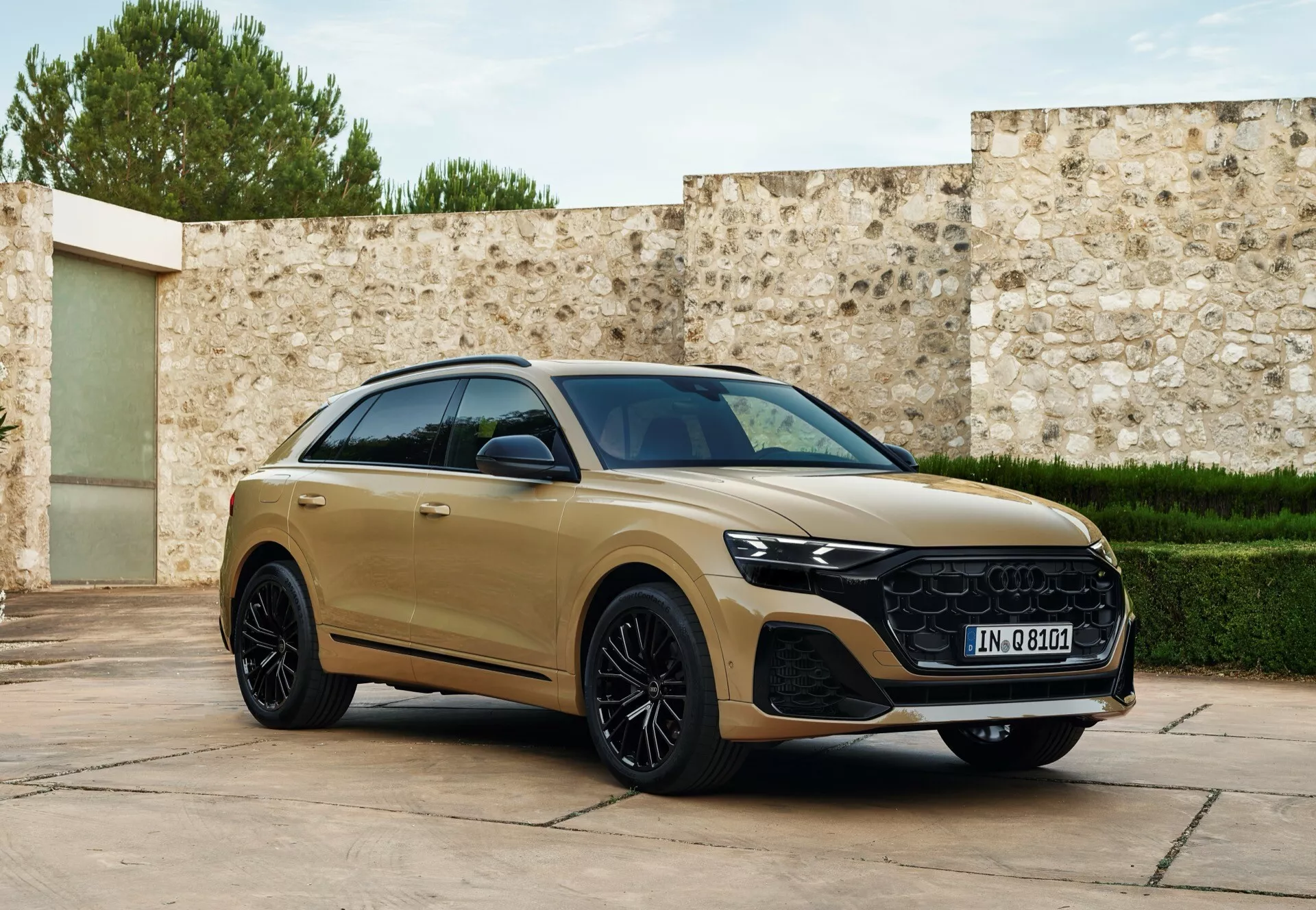 Tričetrtinski sprednji pogled na Audi Q8 2023 (4M-facelift) v zlati barvi, ki prikazuje njegov aerodinamični bočni profil, značilno masko, LED žaromete in luksuzna lita kolesa, parkirano z rahlim zasukom proti kameri na prefinjenem ozadju