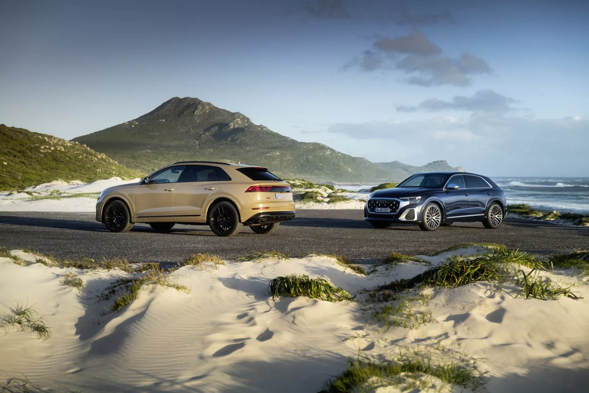 Modeli Audi Q8 (4M-facelift) leta 2023, predstavljeni na peščeni plaži, z zlatim športnim SUV na levi v pogledu zadaj treh četrtin in s srebrnim tradicionalnim SUV na desni v sprednjem pogledu treh četrtin, proti travnatim sipinam, goram in delno oblačnem nebu