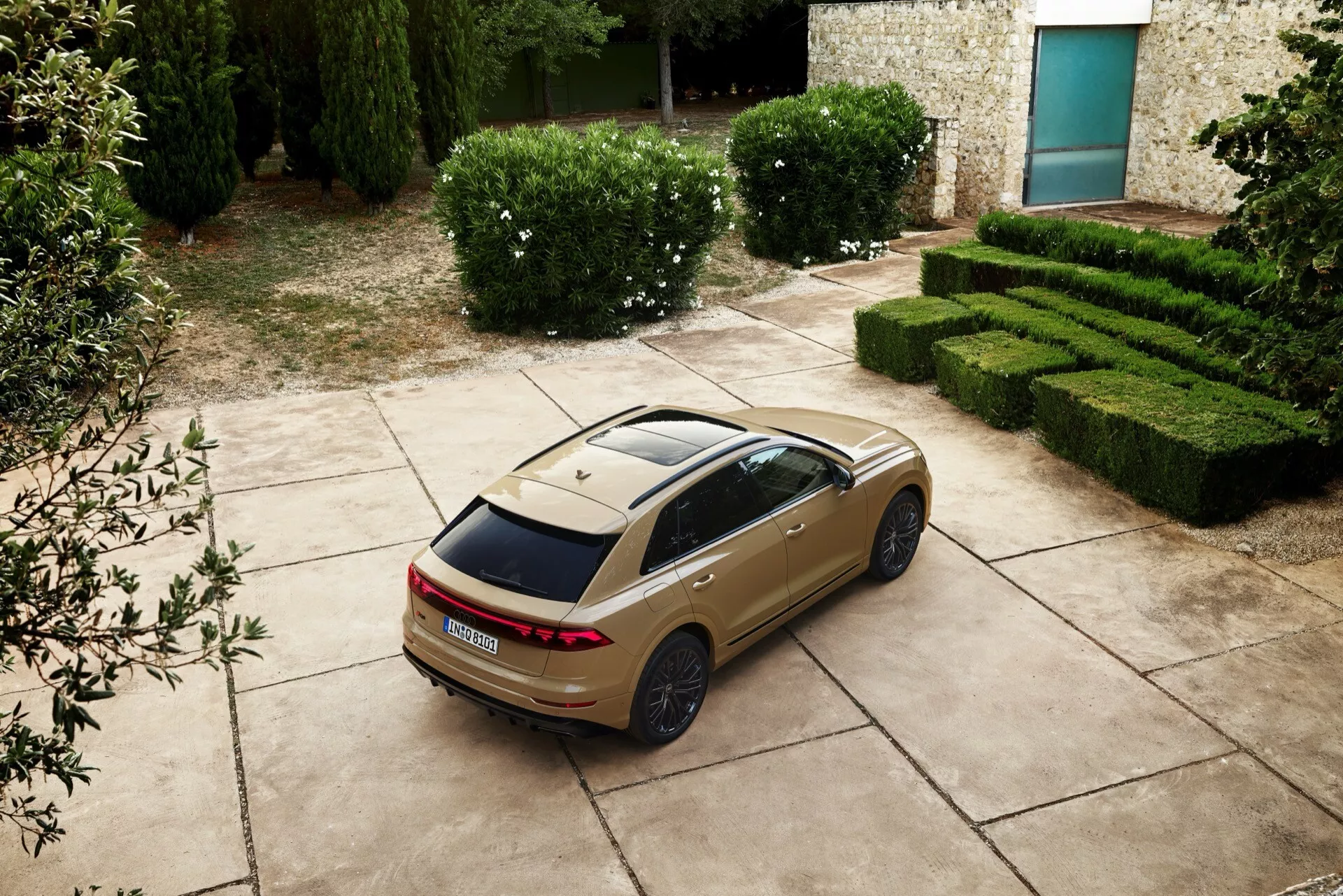 Sedan Audi Q8 2023 (4M-facelift) iz visokega kota, zadnji tričetrtinski pogled prikazuje LED zadnje luči, panoramsko strešno okno in športna platišča, parkiran z idiličnim ozadjem živih mej in kamnite zgradbe