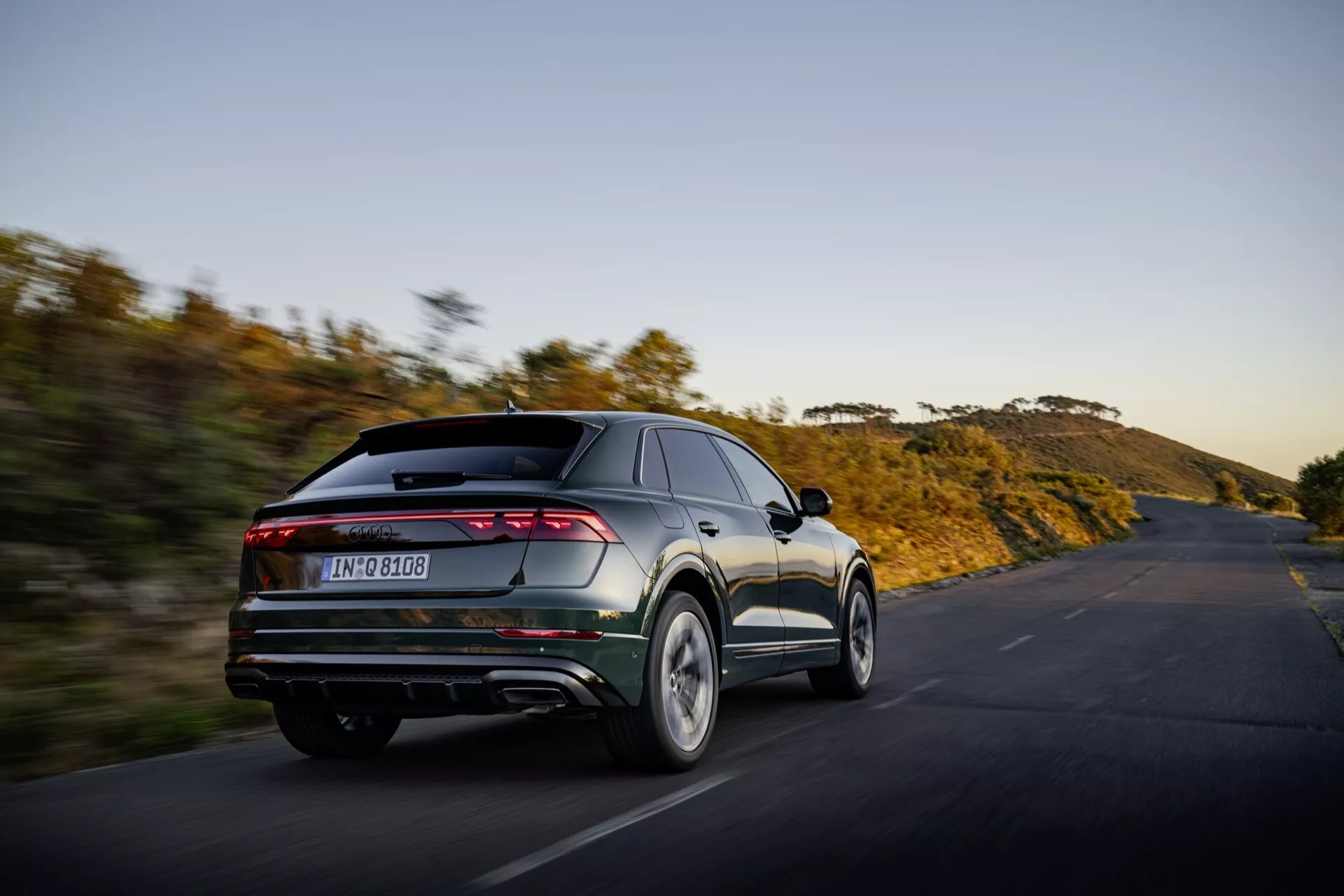 Audi Q8 2023 (4M-Facelift) v gibanju, posnet iz kota treh četrtin zadaj pokazuje sodobne značilnosti oblikovanja vozila vključno z elegantnimi, v celoti širokimi horizontalnimi zadnjimi lučmi, dvojnimi izpušnimi cevmi, zadnjim difuzorjem in integriranim spojlerjem na ozadju naravne pokrajine na odprti cesti ob sončnem vzhodu ali zahodu, ki poudarja njegovo usmerjenost k zmogljivosti in napredni estetiki.