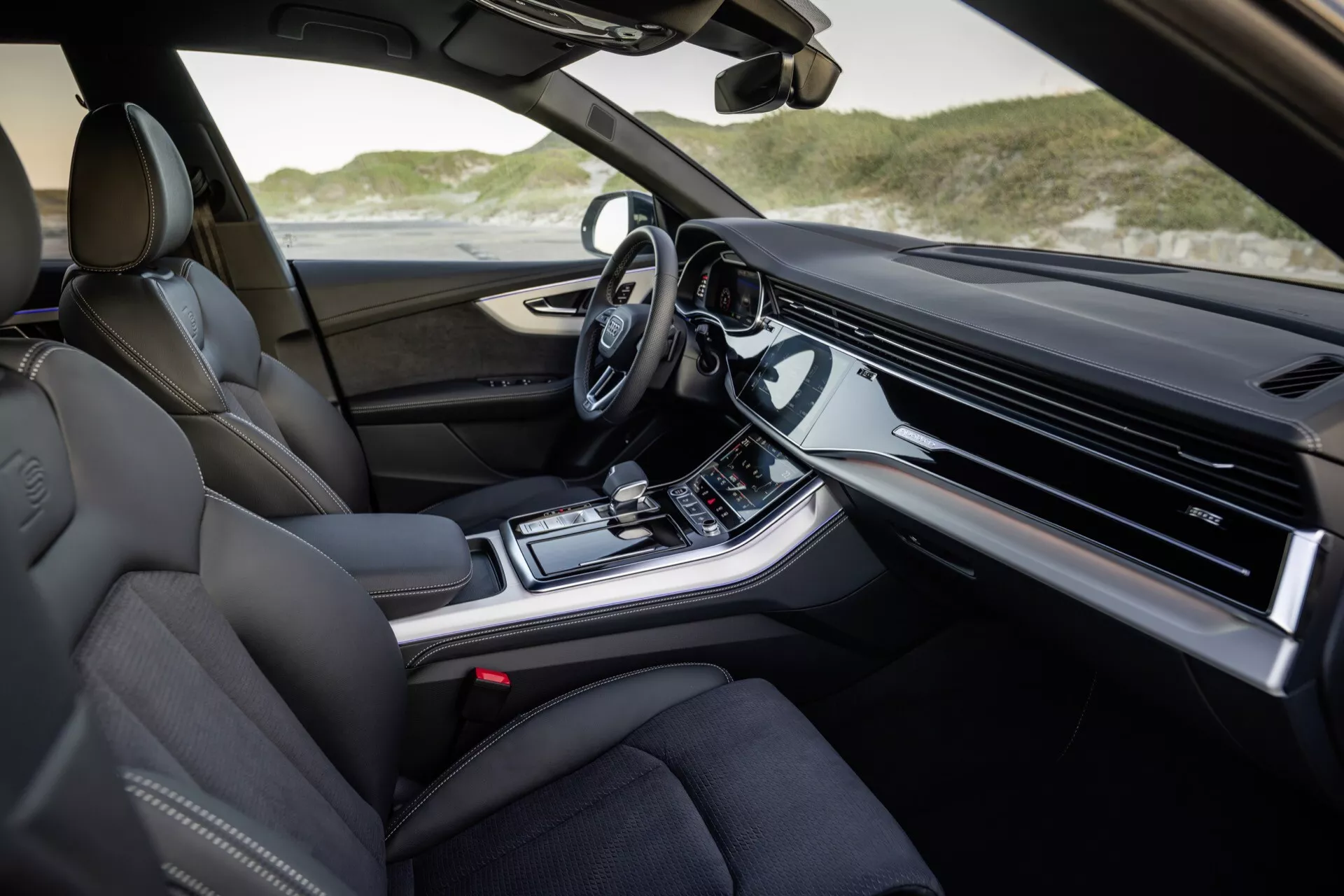 Pogled na notranjost Audi Q8 2023 (4M-Facelift) s strani sopotnika s prestižnimi usnjenimi sedeži, napredno armaturno ploščo z integriranim zaslonom za infotainment, sredinsko konzolo z nadzorom klime in izbiro voznega načina, moderno avtomatsko prestavno ročico in volanom, opremljenim s tehnologijo