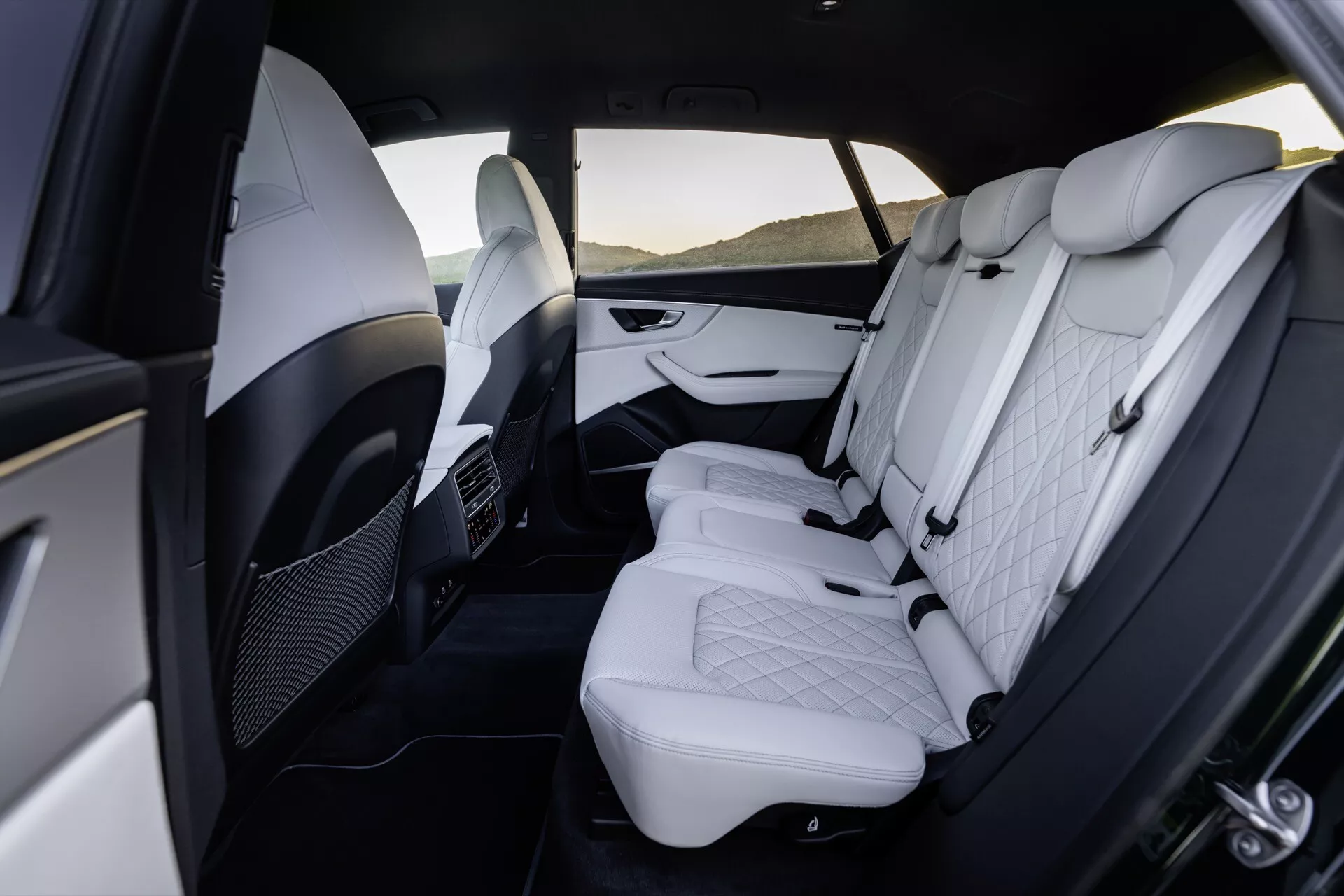 Pogled na luksuzni notranji zadnji del Audi Q8 2023 (4M Facelift), ki prikazuje premium belo usnje z vzorcem prešitih diamantov, nastavljive vzglavnike, dvobarvne plošče vrat z kromiranimi poudarki, zložljivo naslonjalo za roke, prezračevalne odprtine za zadnje potnike in prostorno legroom.