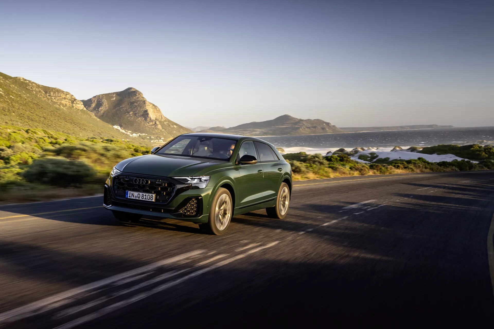 Audi Q8 4M-Facelift 2023 vozi po obalni cesti, poudarjen s svojim aerodinamičnim dizajnom, LED žarometi in emblemom proizvajalca na rešetki, posnet s sprednjim kotom treh četrtin, ki prikazuje luksuzni SUV v gibanju proti slikovitemu ozadju