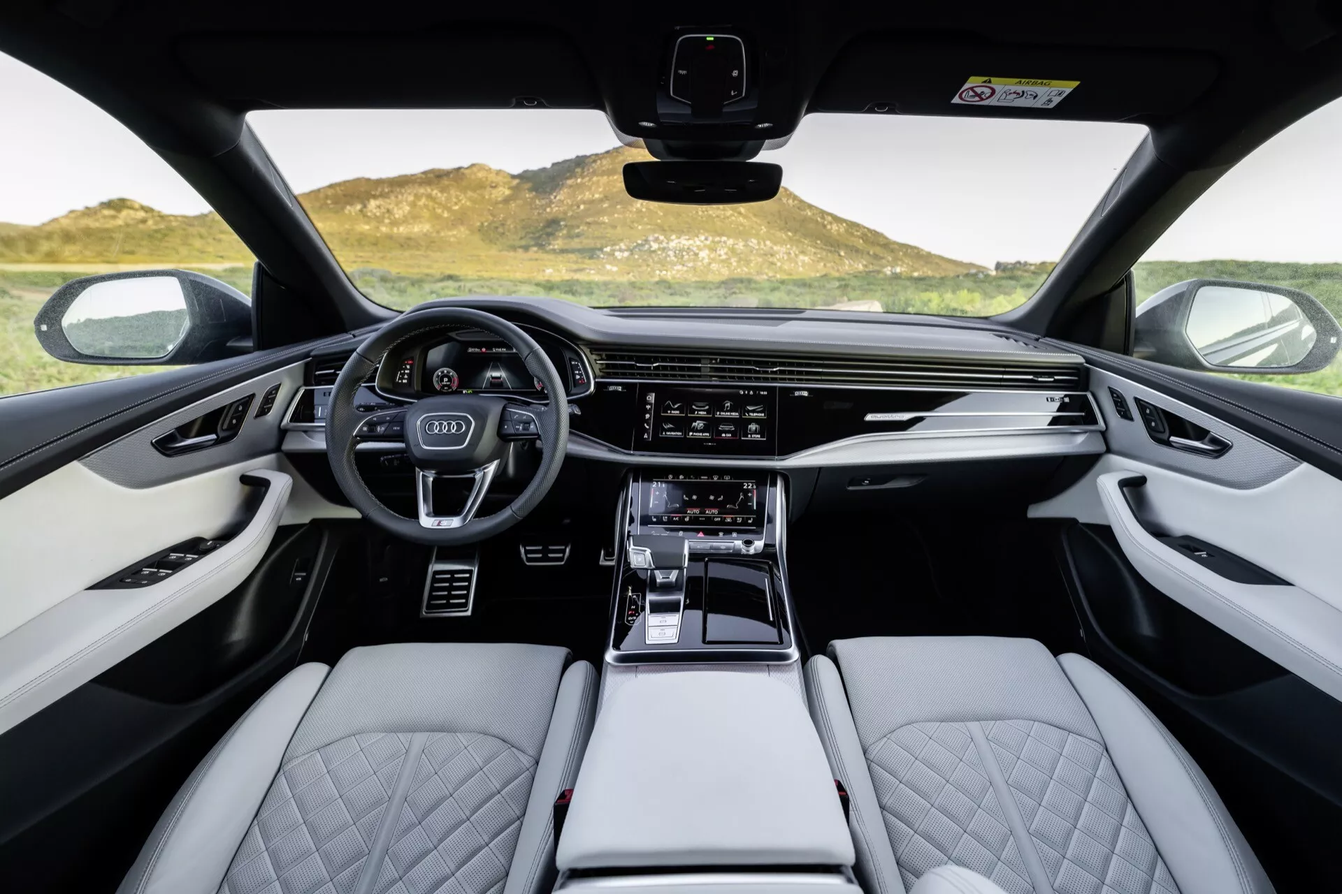 Notranjost Audija Q8 2023 (4M-Facelift), pogled na sprednjo kabino iz perspektive prve osebe s poudarkom na športnem multifunkcijskem volanu, digitalnem instrumentnem zaslonu, širokem zaslonu za infotainment, sedežih iz svetlega usnja s prešitim vzorcem diamanta in detajlih iz visokokakovostnih materialov.