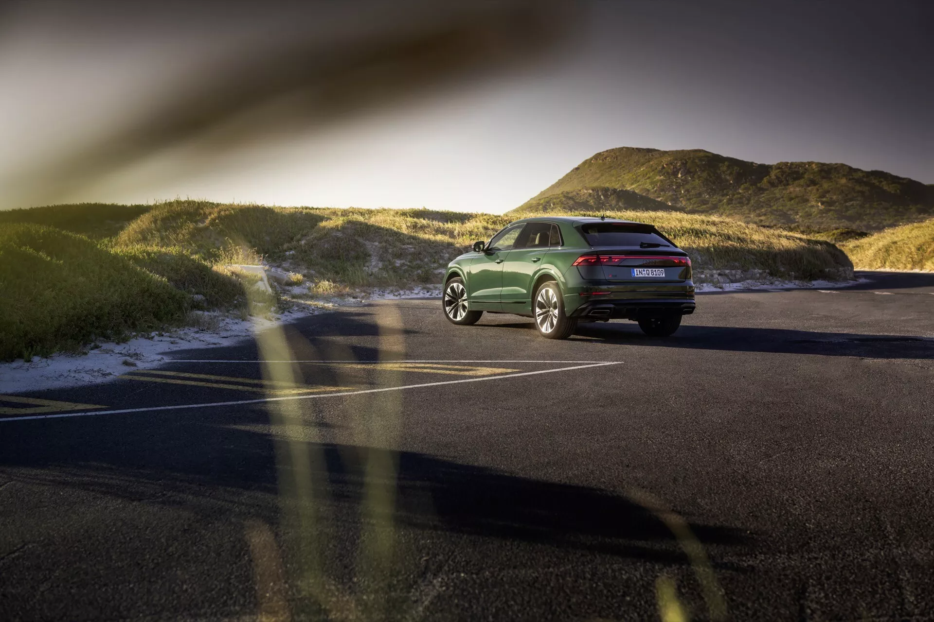 Zadnji tristranski pogled na Audi Q8 (4M-Facelift) 2023 na asfaltnem parkirišču ob sončnem zahodu, ki poudarja njegovo držo, dizajnerske linije, LED zadnje luči, zlitinska kolesa in poliran zeleni zaključek z naravno pokrajino v ozadju.