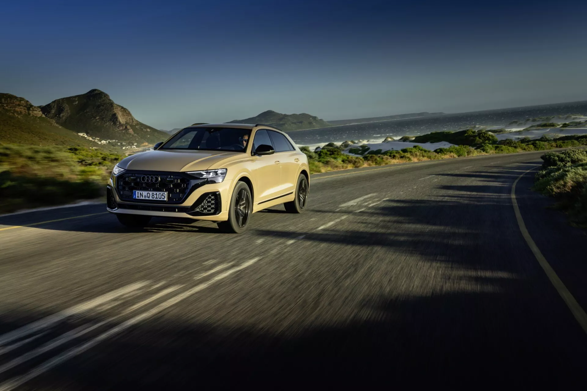 Audi Q8 4M facelift 2023 v gibanju na obalni cesti, ki poudarja napredne LED žaromete luksuznega avtomobila, izstopajočo masko, aerodinamičen pokrov in dinamično upravljanje, postavljen proti ozadju gora in oceana s poudarkom na oblikovnih in tehnoloških lastnostih.