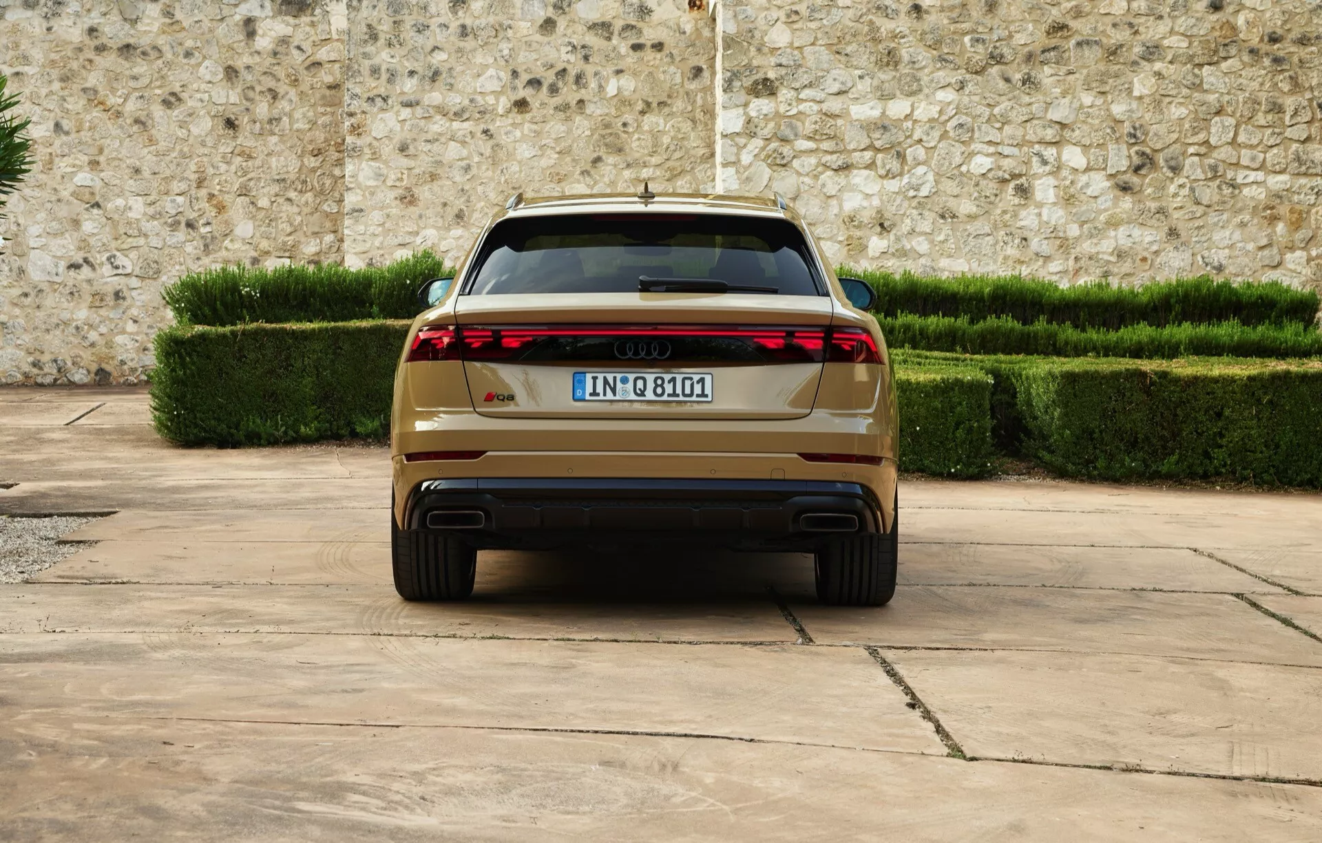 Audi Q8 2023 (4M-Facelift) prikazuje zadnji pogled z modernimi LED zadnjimi lučmi, dvojnimi izpuhi, evropsko registrsko tablico, parkiran na tlakovani površini proti ozadju kamnitega zidu in urejenih zelenih živih mej, kar poudarja luksuz in napredne oblikovalske značilnosti