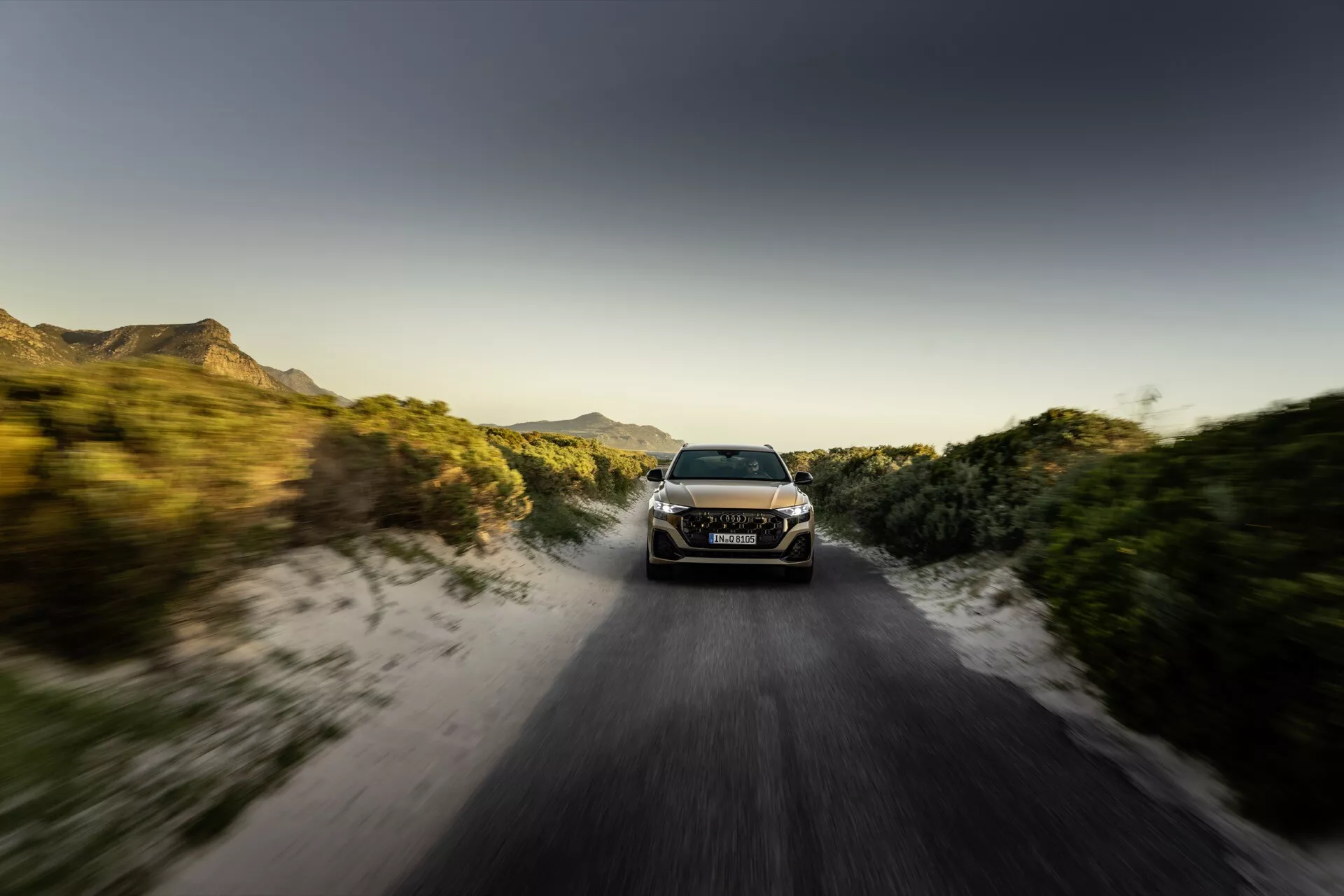 Audi Q8 2023 (4M-Facelift) v dinamičnem gibanju po neasfaltirani cesti ob sončnem vzhodu ali zahodu z učinkom zamegljenega gibanja, ki prikazuje svoje LED žaromete, elegantno aerodinamično obliko in terensko sposobnost sredi podeželskega okolja