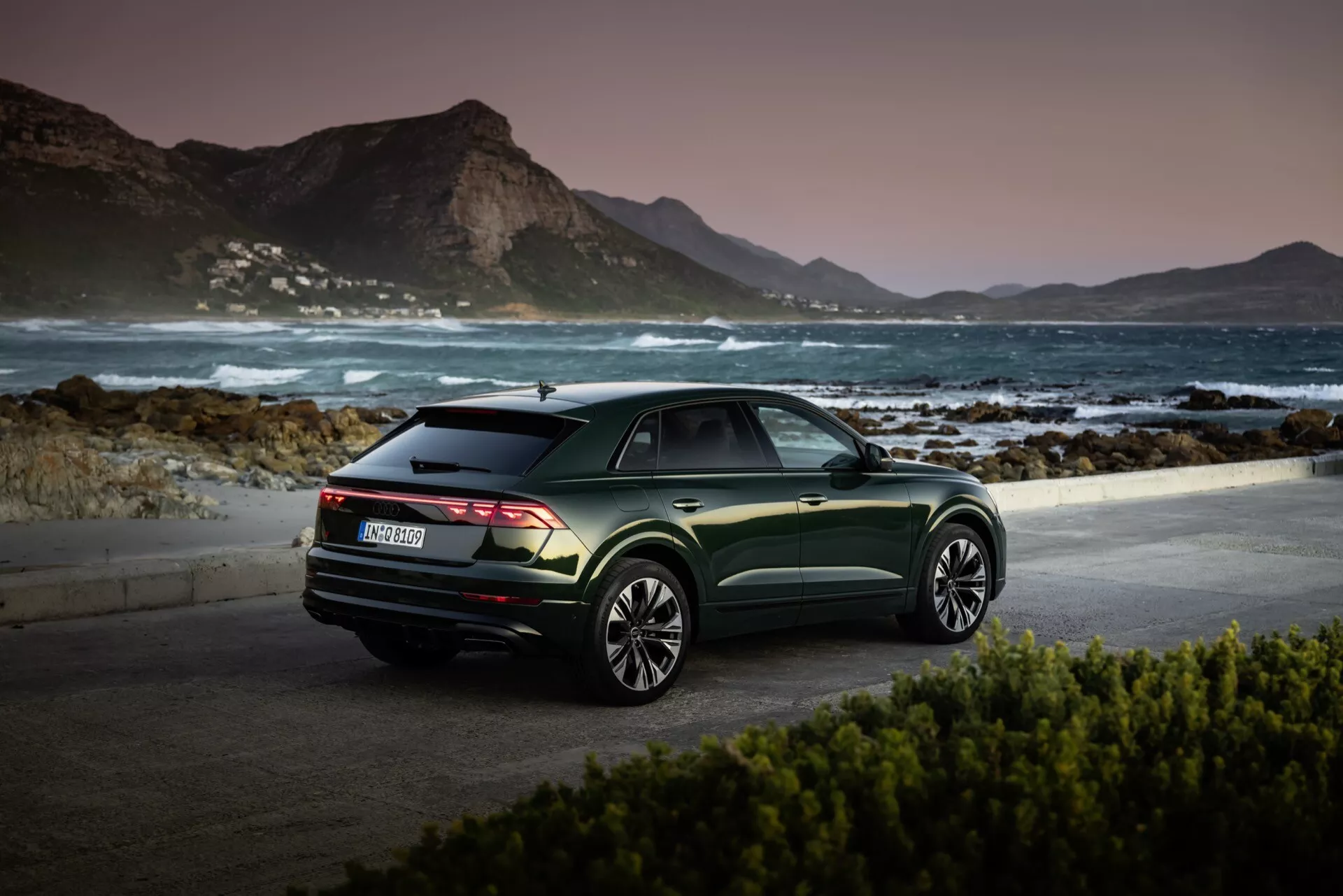 Audi Q8 2023 (4M-Facelift) parkiran na slikoviti obalni lokaciji ob svitu ali mraku, ki prikazuje svoj pogled od zadaj, temno zeleno kovinsko barvo in večkraka lita kolesa proti mirni gorski kulisi