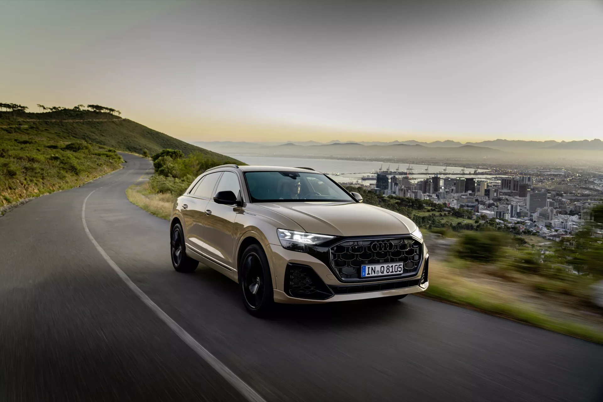 Luksuzni križanec SUV Audi Q8 2023 (4M-Facelift) v gibanju na ovinkasti cesti, prikazuje športno oblikovanje, LED žaromete, velika platišča iz zlitine in panoramsko streho, proti ozadju mesta in gora v mehki jutranji ali popoldanski svetlobi.