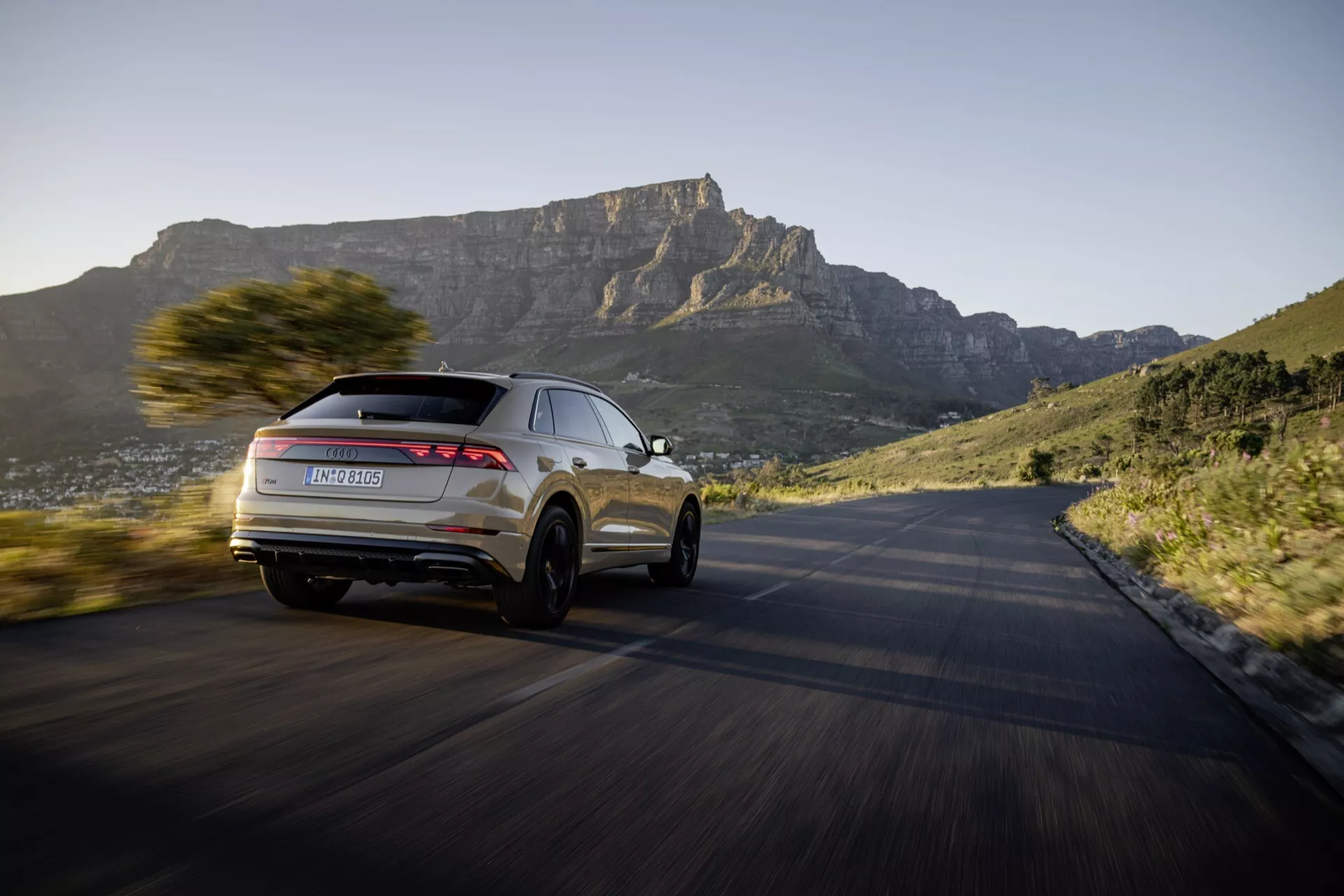 Model Audi Q8 4M facelift 2023 v dinamičnem gibanju na ovinkasti cesti, poudarjajoč njegov sodoben dizajn SUV in napredne funkcije iz perspektive treh četrtin od zadaj, z idiličnim gorskim ozadjem ob sončnem zahodu