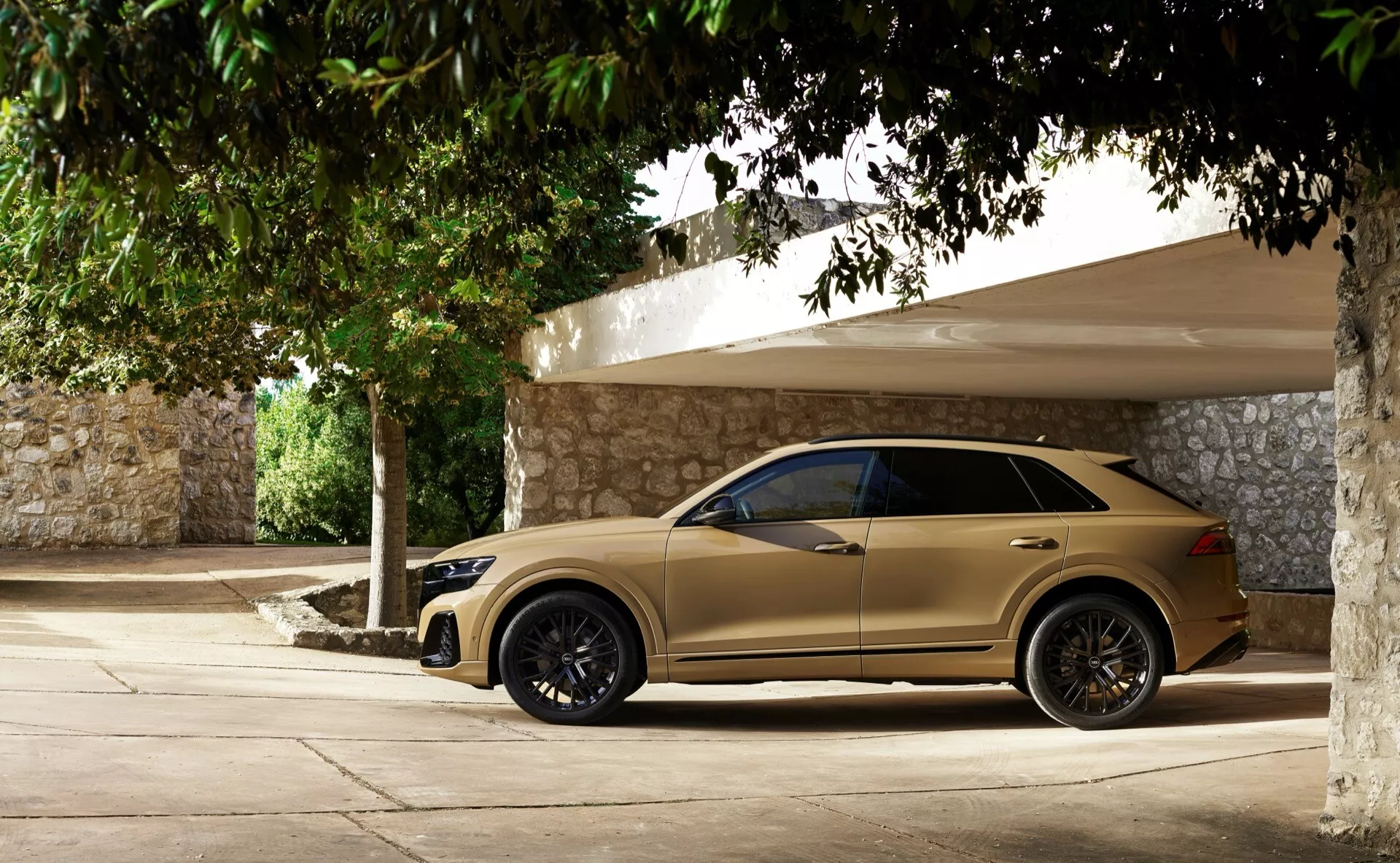 Audi Q8 2023 (4M-Facelift) v bež/zlati barvi, prikazuje celoten stranski pogled s temnimi litimi platišči, ogledali z integriranimi smerniki, zatemnjenimi okni, vratnimi kljukami in poševno linijo strehe, parkiran v senci na sončen dan