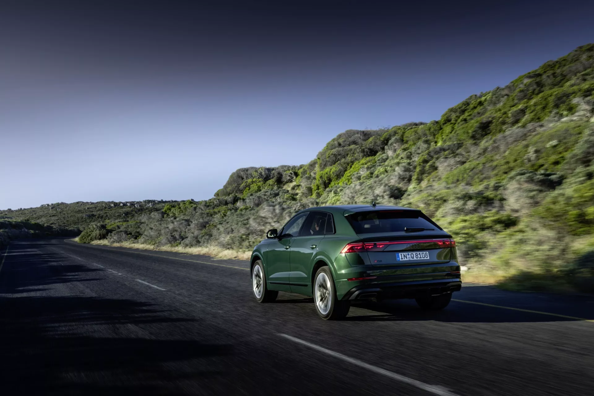 Audi Q8 2023 (4M-Facelift) v dinamičnem gibanju na obalni ali gorski cesti, ki prikazuje svoj slog sportback, elegantne zadnje luči in sofisticirana lita kolesa proti dramatični pokrajini