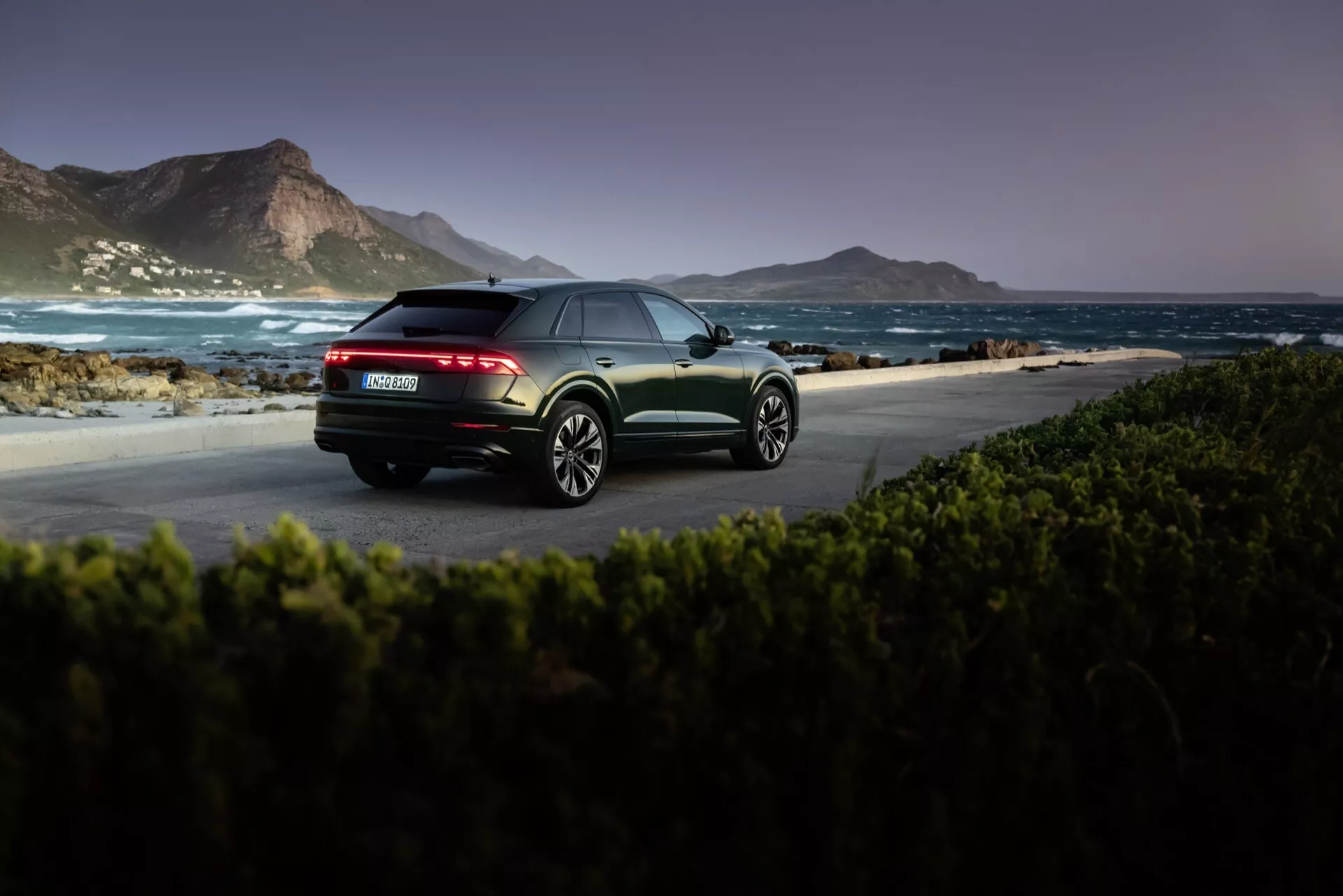 Audi Q8 4M-Facelift 2023 v kovinski barvi, posnet ob zori ali mraku v naravnem obalnem okolju, poudarja pogled na zadnji del vozila z LED zadnjimi lučmi, dvojnimi izpuhi in večkraki lita platišča.