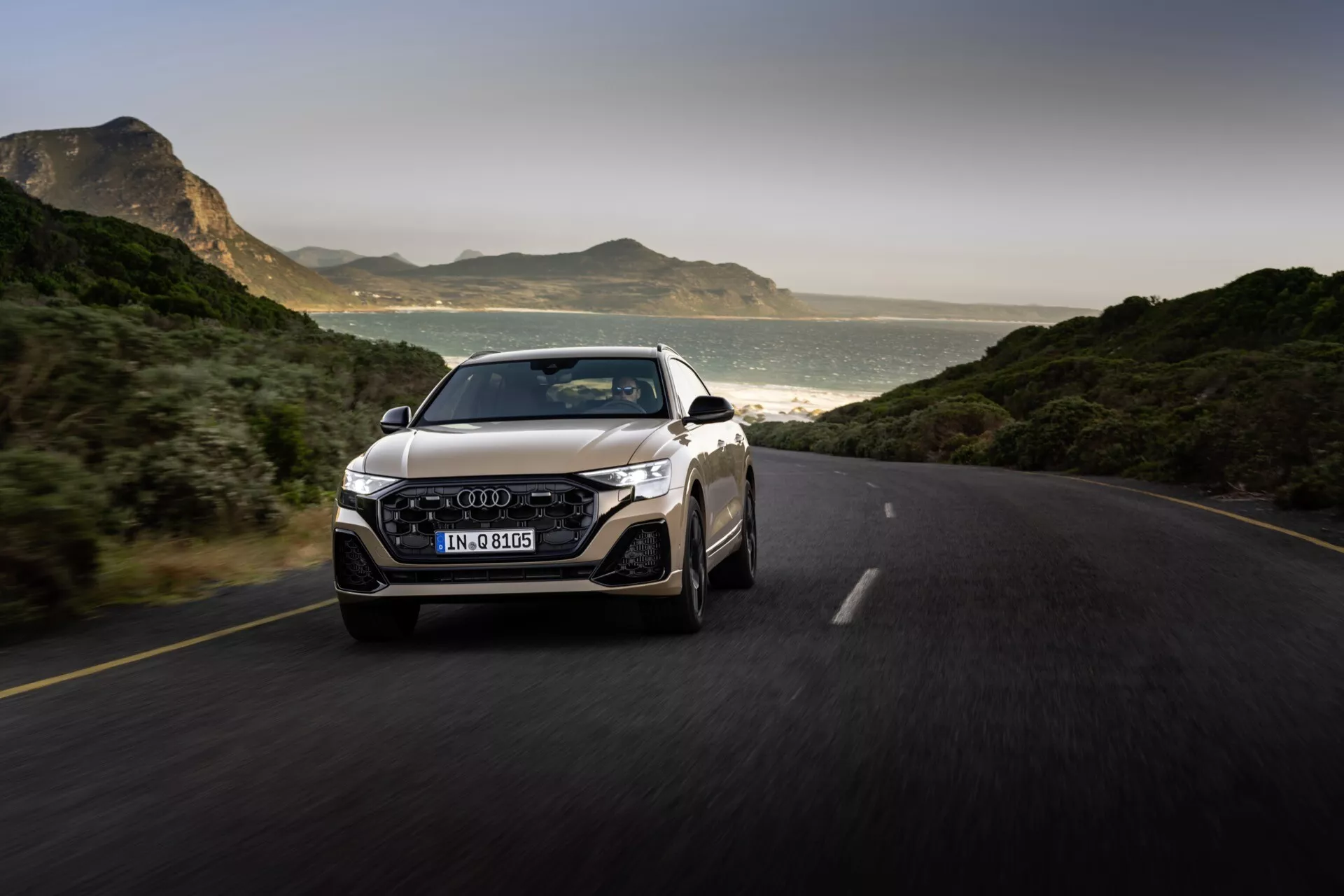 Audi Q8 2023 (4M-Facelift) ujeto v gibanju na asfaltirani cesti, ki prikazuje svojo dinamično zasnovo iz sprednjega tričetrtinskega pogleda, poudarjajoč žar, LED žaromete, odbijač in emblema na žaru, z idiličnim ozadjem gora in obale pod naravno svetlobo