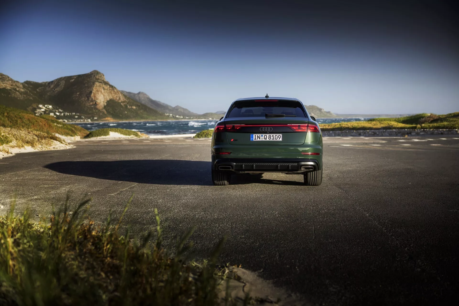 Zadnji pogled na model Audi Q8 4M Facelift 2023, ki prikazuje LED zadnje luči, kromirane poudarke in dvojno izpušno cev na slikoviti obalni cesti
