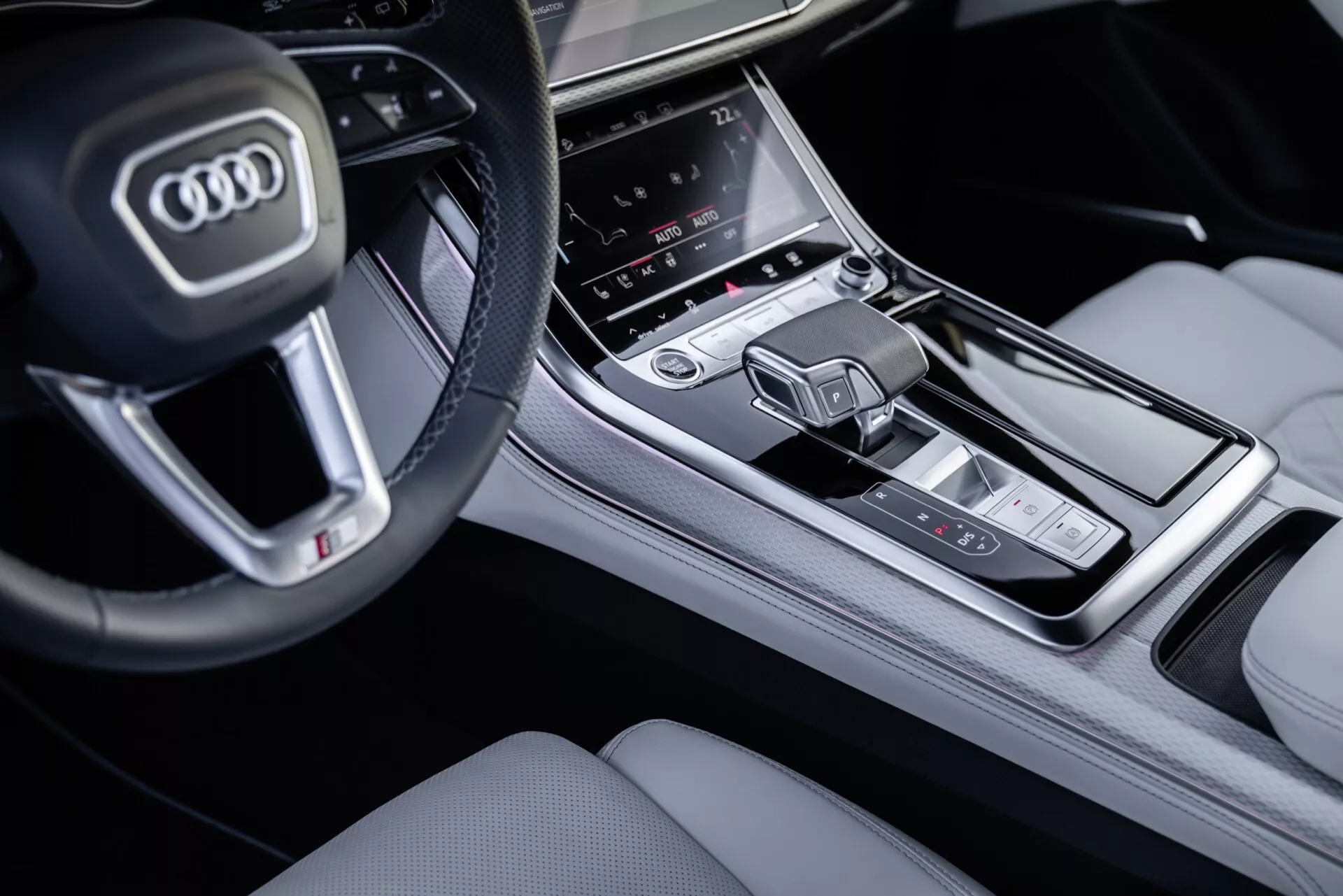 Podroben pogled na sredinsko konzolo in del volana Audija Q8 2023 (4M-Facelift) z vidika sovoznika, izpostavljajoč gumbe na multifunkcijskem volanu, centralni zaslon na dotik za infozabavni sistem, območje za nadzor klime in moderni izbirnik prestav v premijski notranji opremi.