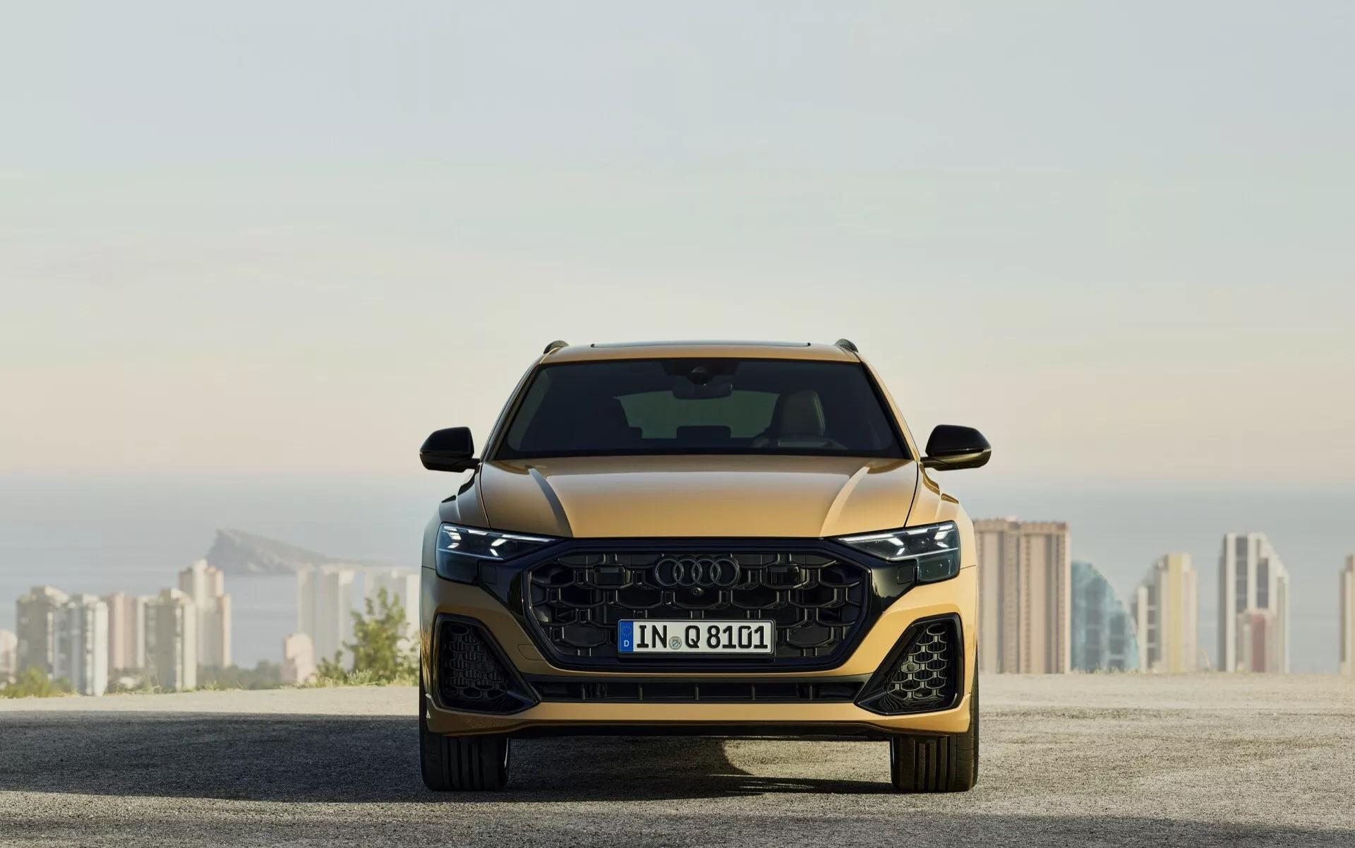 Celoten čelni pogled na Audi Q8 (4M-Facelift) 2023, ki prikazuje njegov aerodinamičen dizajn, veliko masko z logotipom štirih obročev, vitke LED svetlobne enote in integrirane napredne sisteme pomoči vozniku, na ozadju zabrisane mestne pokrajine