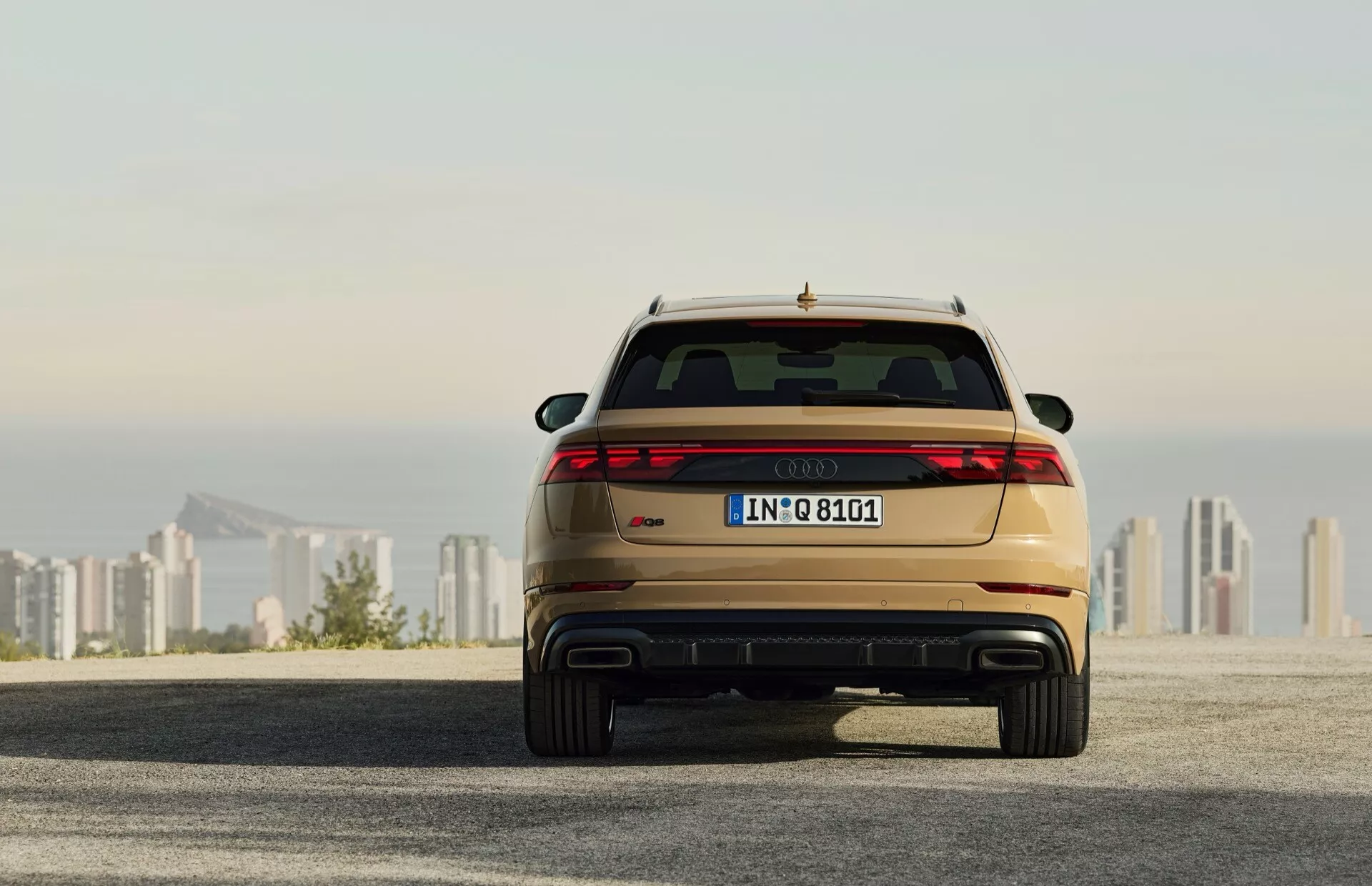 Zadnji pogled na Audi Q8 2023 (4M-Facelift), ki prikazuje LED zadnje luči, dvojni izpuh in kovinsko barvo, parkiran na asfaltni površini z mestno obzorje v ozadju ob sončnem vzhodu/zahodu