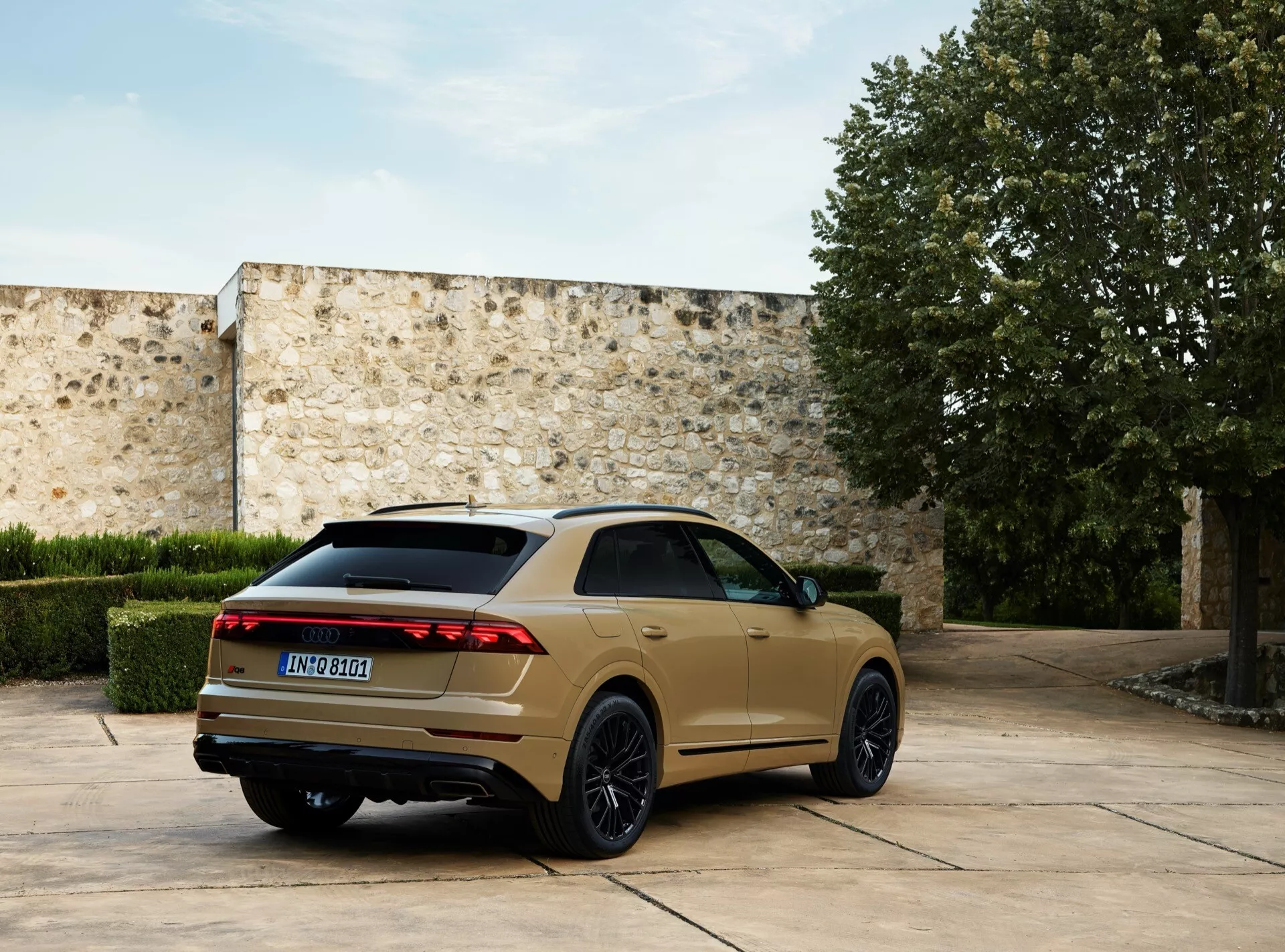 Model Audi Q8 4M facelift 2023 parkiran zunaj, posnet z zadnjega kota tri četrtine, ki poudarja njegove LED zadnje luči, temna večkraka lita kolesa, padajočo streho in dvojne izpušne cevi proti ozadju dvorišča iz kamna
