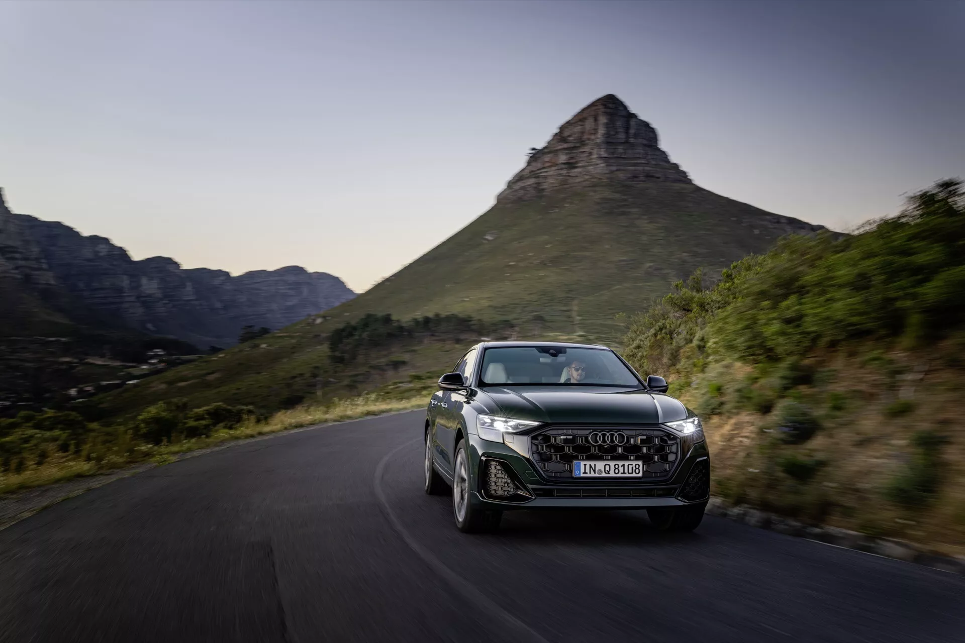 Audi Q8 2023 (4M-Facelift) v dinamičnem gibanju na slikoviti vijugasti cesti, ki prikazuje svojo aerodinamično obliko, LED žaromete, značilna lita kolesa in integrirane zračne reže proti idilični gorski kulisi v poznem popoldnevu