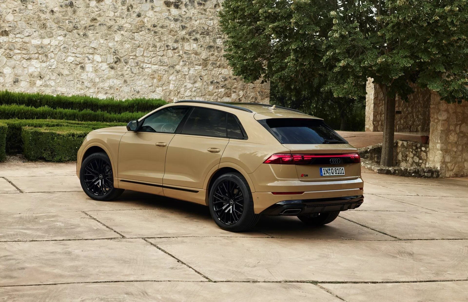 Audi Q8 2023 (4M-Facelift) prikazan z zadnjega pogleda za tri četrtine, ki prikazuje velika črna platišča z večimi kraki, kovinsko zlato-bež barvo, podolgovate horizontalne LED zadnje luči, zadnji spojler in dvojni izpušni sistem pred zgodovinskim kamnitim zidom