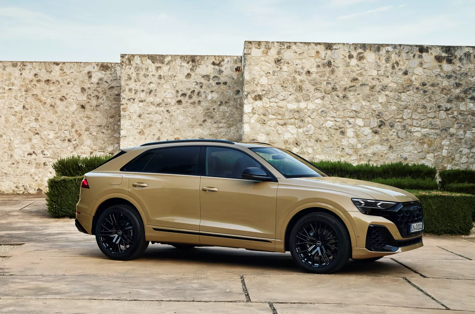 Trikvartni sprednji pogled na moderni Audi Q8 2023 (4M-Facelift) v mat zlati ali bež barvi z veliko rešetko s satovjem, LED žarometi, športnim sprednjim odbijačem, črnimi zlitinskimi kolesi, pnevmatikami z nizkim profilom, vgradnimi kljukami vrat, temno obarvanimi okni, spuščajočo se strešno linijo v slogu coupé, mišičasto bočno linijo z angularnimi zadnjimi lučmi in strešnim spojlerjem, postavljeno proti teksturirani steni, kar nakazuje na zgodovinsko ali sodobno okolje.