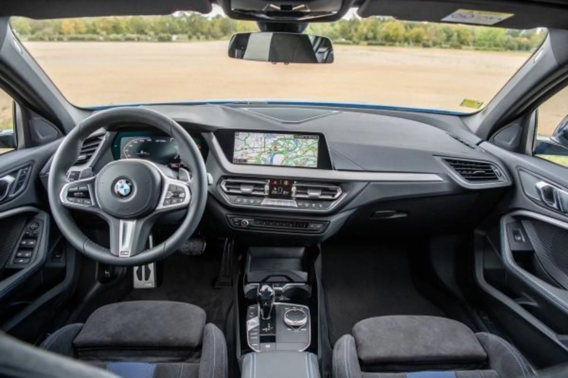 Notranjost BMW serije 1 M Performance 2019 iz perspektive zadnjega sedeža prikazuje armaturno ploščo, sredinsko konzolo in volan z nameščenimi kontrolniki, zaslon na dotik za infotainment sistem, ki prikazuje GPS navigacijo, zračnike, kontrole klime, avtomatsko prestavno ročico, vrtljiv gumb in sledilno ploščico na sredinski konzoli, vrhunsko oblazinjenje in svetlo, dobro osvetljeno kabino
