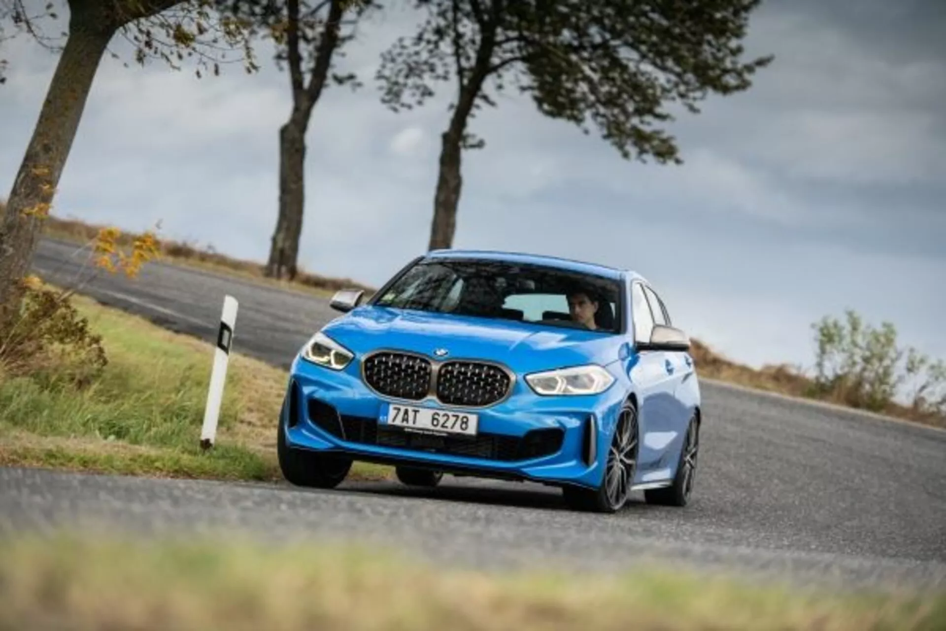 Modri športni sedan BMW serije 1 M Performance letnik 2019, posnet iz nizkega in kotnega stransko-sprednjega zornega kota, ki poudarja njegov agresiven sprednji del z ikoničnimi ledvičnimi režami, elegantnimi LED žarometi, velikimi zračnimi režami za hlajenje zmogljivosti, nizkim športnim vzmetenjem, v gibanju okoli ovinka, ki kaže dinamično vožnjo, z aerodinamičnimi stranskimi ogledali, velikimi litoželeznimi kolesi in pnevmatikami z nizkim profilom, ki poudarjajo športni status in zmožnosti oprijema.