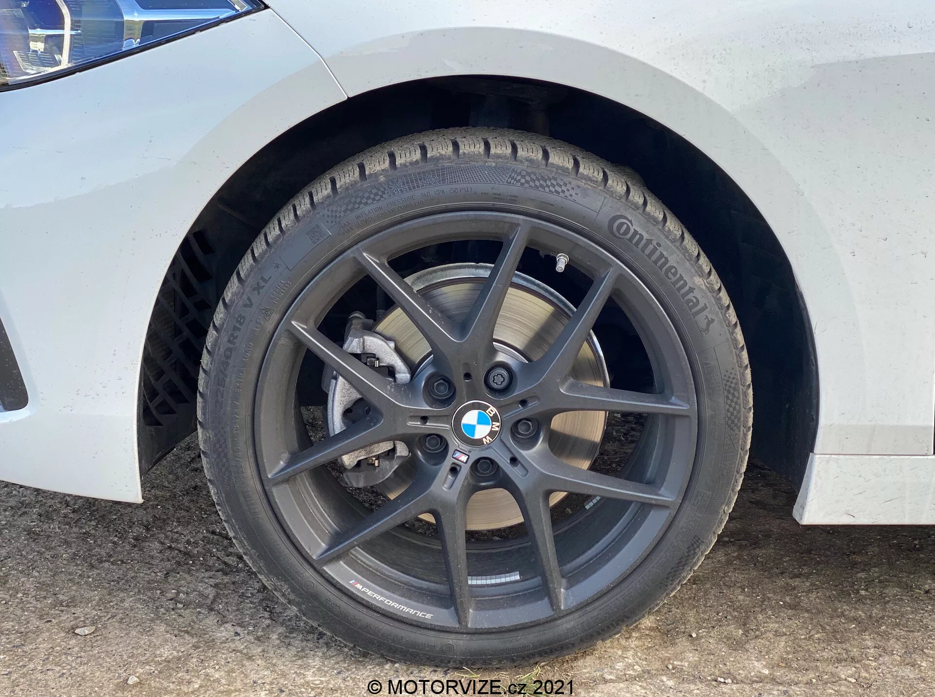 Povečava prednjega kolesa in blatnika vozila BMW 1er-Reihe 2019 (BMW-116i). Perspektiva na ravni tal poudarja temno sivo petkrako platišče iz lahke litine z BMW-emblemom, blagovno znamko pnevmatike Continental z vidnim profilom, ventilirano zavorno ploščo in srebrno enobatno zavorno čeljust. Bela barva s prahom in vodnimi madeži kaže na uporabo.