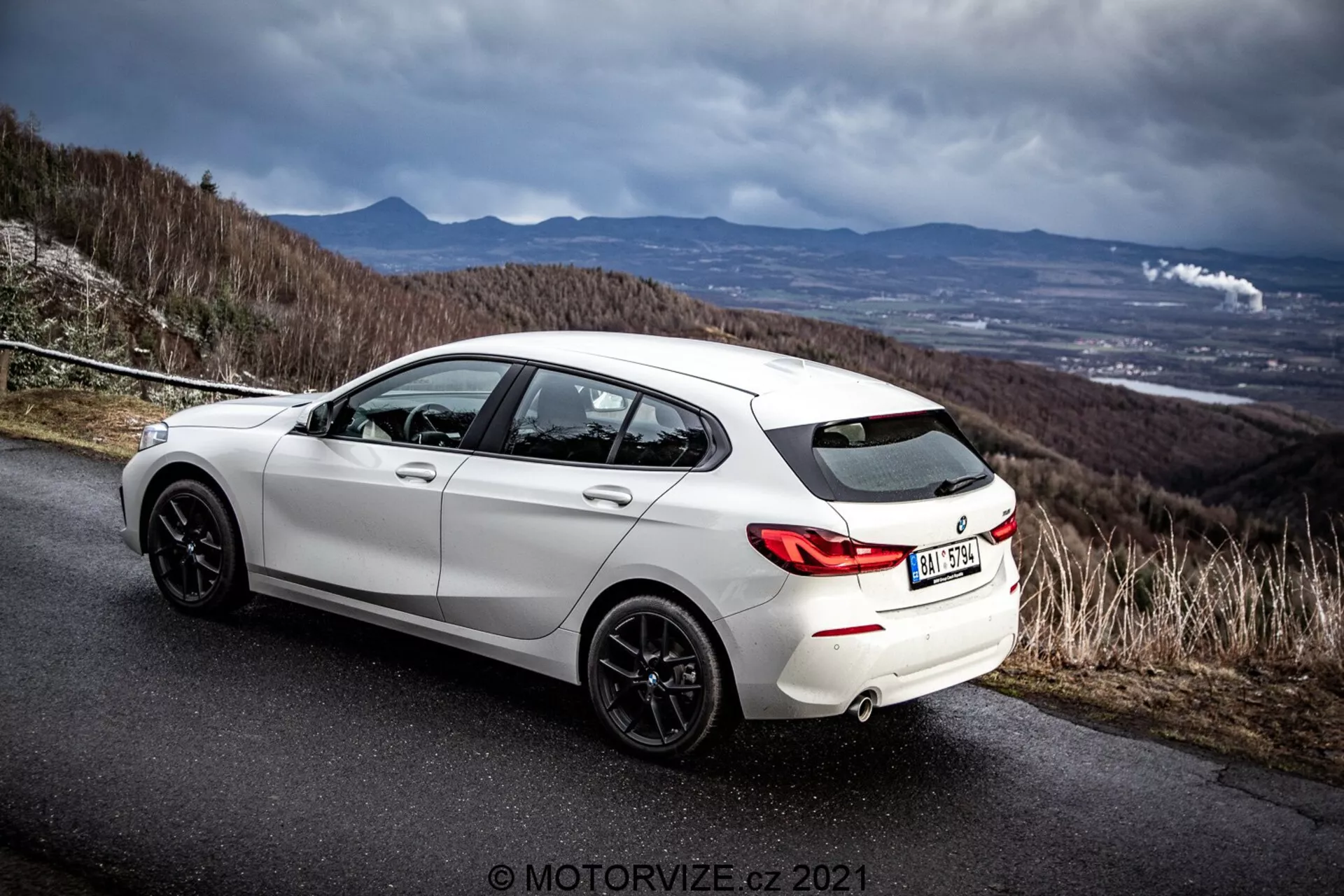 Poševni zadnji stranski pogled na bel hatchback BMW 116i iz leta 2019 na asfaltirani cesti z dramatičnim oblačnim nebom. Slika poudarja zadnjo zasnovo vozila, zadnje obrobe koles, temno barvana platišča, dvojni izpušni sistem in stranski profil, vključno z zadnjimi potniškimi vrati in vrati voznika. Okolje nakazuje hribovsko ali gorsko cesto in poudarja športne in dinamične lastnosti avtomobila. Navzoč je vodoznak fotografa 'MOTORWIZE.cz 2021'.