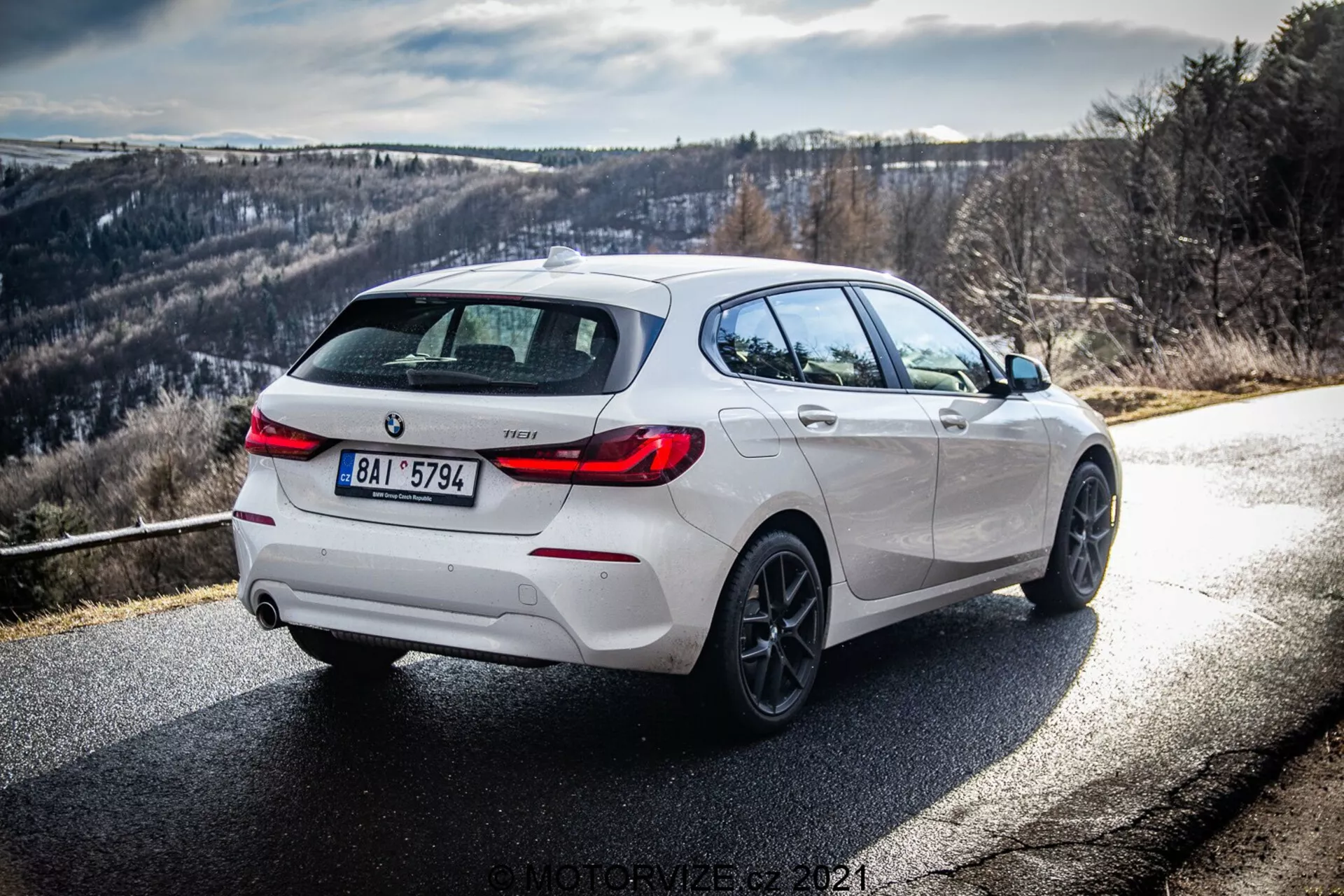 Dinamičen zadnji pogled treh četrtin belega BMW 116i 2019, ki prikazuje desno stran in zadek podnevi s čistimi sončnimi žarki in sencami. Sliko poudarjajo oblikovne podrobnosti, vključno s črnimi poudarki na zadnjem difuzorju, platiščih in ohišju stranskih ogledal, dopolnjene z lita platišča in dvojni izpušni sistem, kar nakazuje na športno različico. Cesta se zavije v desno z hribovitim ali goratim ozadjem, idealna za ljubitelje vožnje.