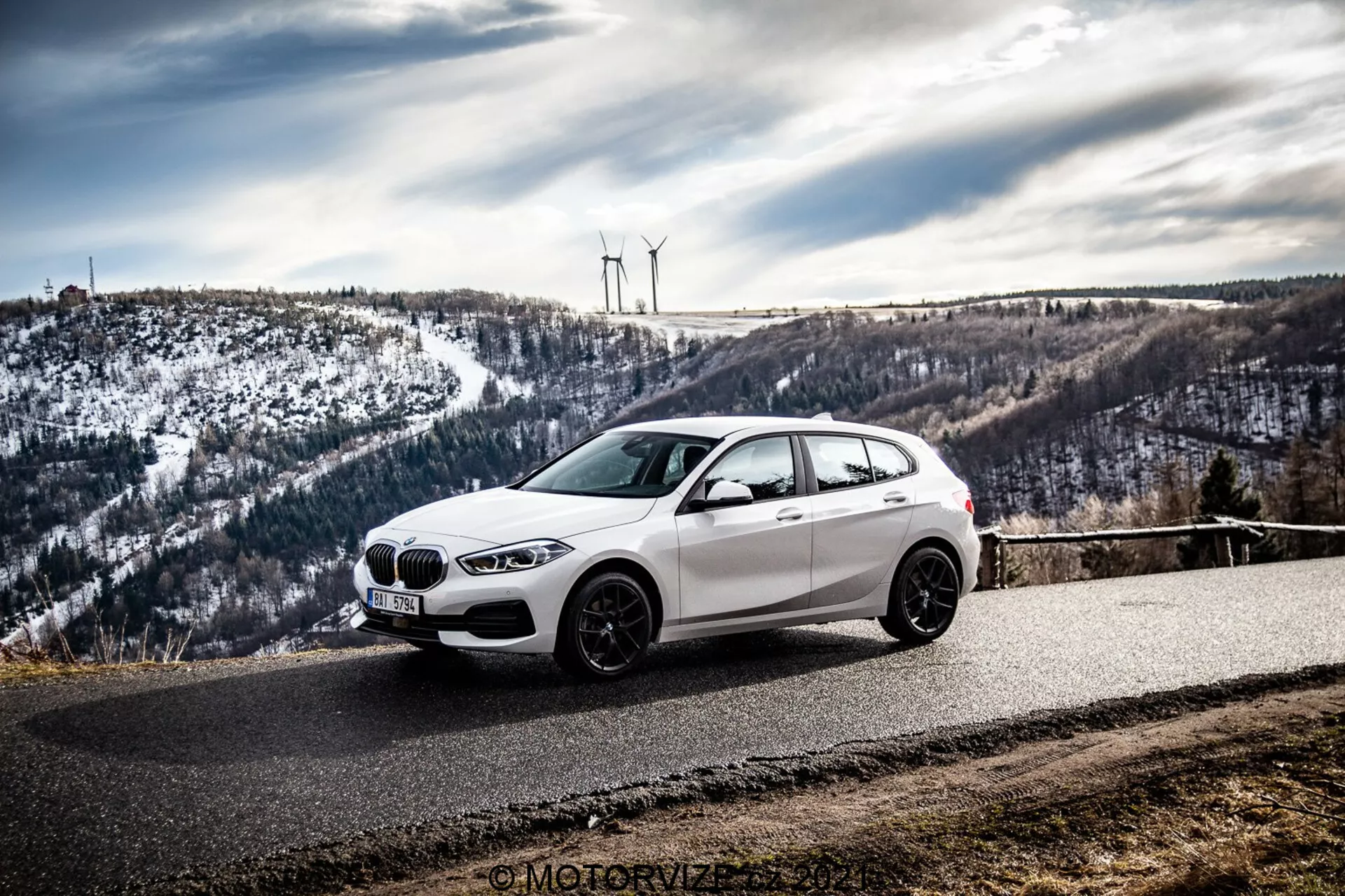 Bel BMW hatchback avtomobil, verjetno BMW 1er-Reihe 2019 (BMW 116i), je parkiran na zaviti asfaltirani cesti, obdana s hribovito, zasneženo pokrajino z vetrnimi turbinami. Fotografija, posneta iz bočno-sprednjega kota, poudarja sprednjo mrežo, žaromete, aerodinamične linije in aluminijasta platišča avtomobila pod oblačnim nebom.