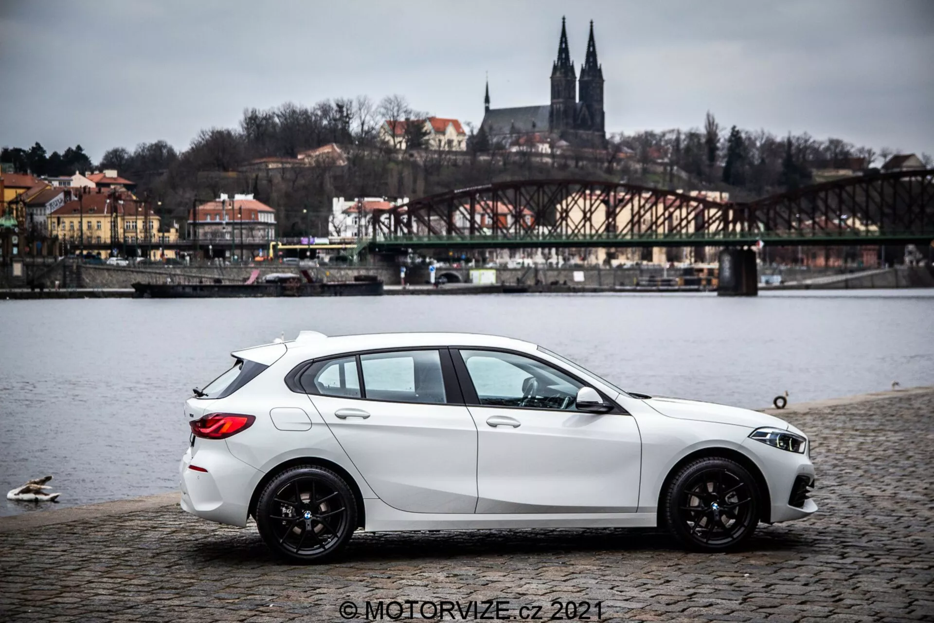 Bel hatchback BMW serije 1 iz leta 2019 (BMW-116i), parkiran na tlakovanem površju v bližini vodnega telesa s racami, prikazan v bočnem profilu z rahlimi zadnjimi kvadranti. Avto ima dinamične oblikovane linije, padajočo strešno linijo, črna večkraka lita platišča ter vidne žaromete in stranska ogledala. V ozadju so most, stavbe na hribu in arhitekturni elementi, ki kažejo na evropsko okolje. Osvetlitev je razpršena pod oblačnim nebom, slika pa vsebuje vodni žig © MOTORVIZE.cz 2021.