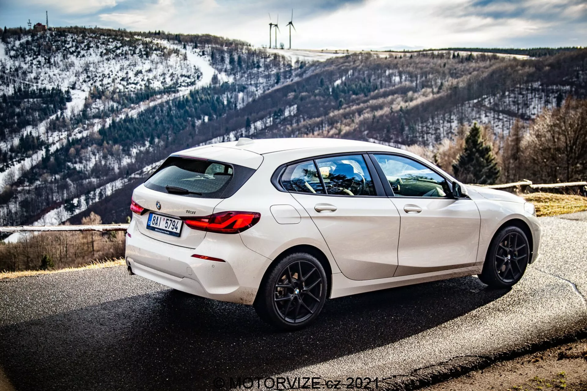 Slika prikazuje bel BMW serije 1 (BMW-116i) model 2019 parkiran ob cesti s slikovitim ozadjem, ki vključuje hribovit, zasnežen pejsaž in vetrne turbine na obzorju. Fotografija je posneta iz zadnjega kota pod kotom treh četrt, ki jasno prikazuje desno stran avtomobila. Pogled poudarja elegantne oblikovalske linije, zadnji spojler, petkraka črna platišča, dvojna izpušna cev in zadnja zatemnjena stekla. Avto ima tudi zadnjega brisalca in vgrajeno strešno antenski shark-fin. Globina polja ohranja avto ostro osredotočeno z naravno dnevno svetlobo, ki poudarja njegovo športno estetiko, medtem ko je ozadje subtilno podrobno.