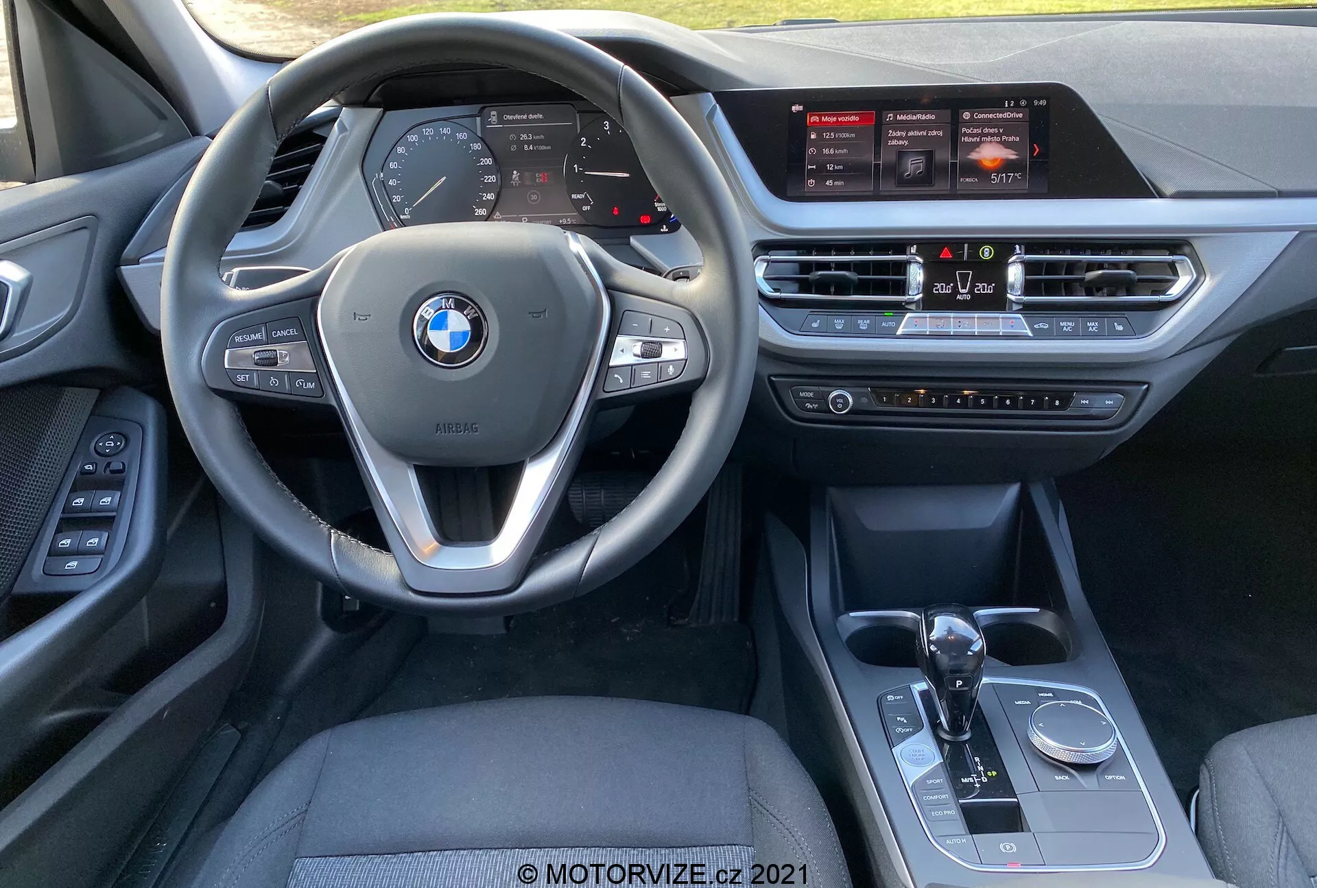 Notranji pogled BMW 1. serije (BMW 116i) 2019 iz perspektive sprednjega potniškega sedeža, ki prikazuje večfunkcijski volan z vgrajenimi kontrolami, digitalno instrumentno ploščo, velik zaslon na dotik za informativno-zabavni sistem, sistem za nadzor klime, samodejno prestavno ročico, izbiro načina vožnje in nadzorni gumb iDrive.