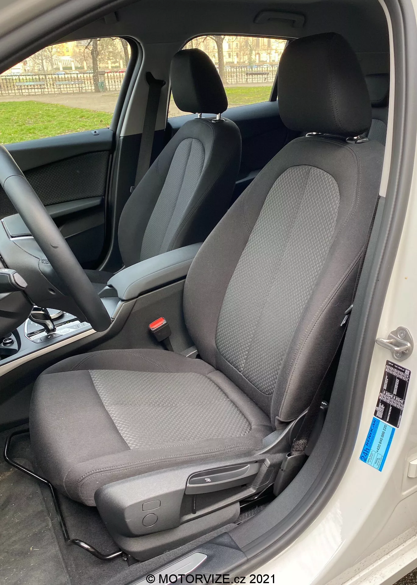 Pogled od blizu na notranjost BMW 1er-Reihe 2019 (BMW-116i), osredotočen na voznikov sedež z modernim dvobarvnim oblikovanjem tkanine, večinoma črne barve z svetlejšimi sivimi vzorci, posnet iz stranskega kota s široko odprtimi vrati vozila. Sedež ima vzglavnik proti svetlobi, kar ustvarja silhuetni učinek okoli robov, in vključuje ročico za ročne nastavitve naprej ali nazaj, pa tudi majhen črn gumb blizu roba notranjosti vozila. Naravna dnevna svetloba poudarja ostro osredotočenost na sedež, z zamegljenim ozadjem, ki prikazuje volan in armaturno ploščo.