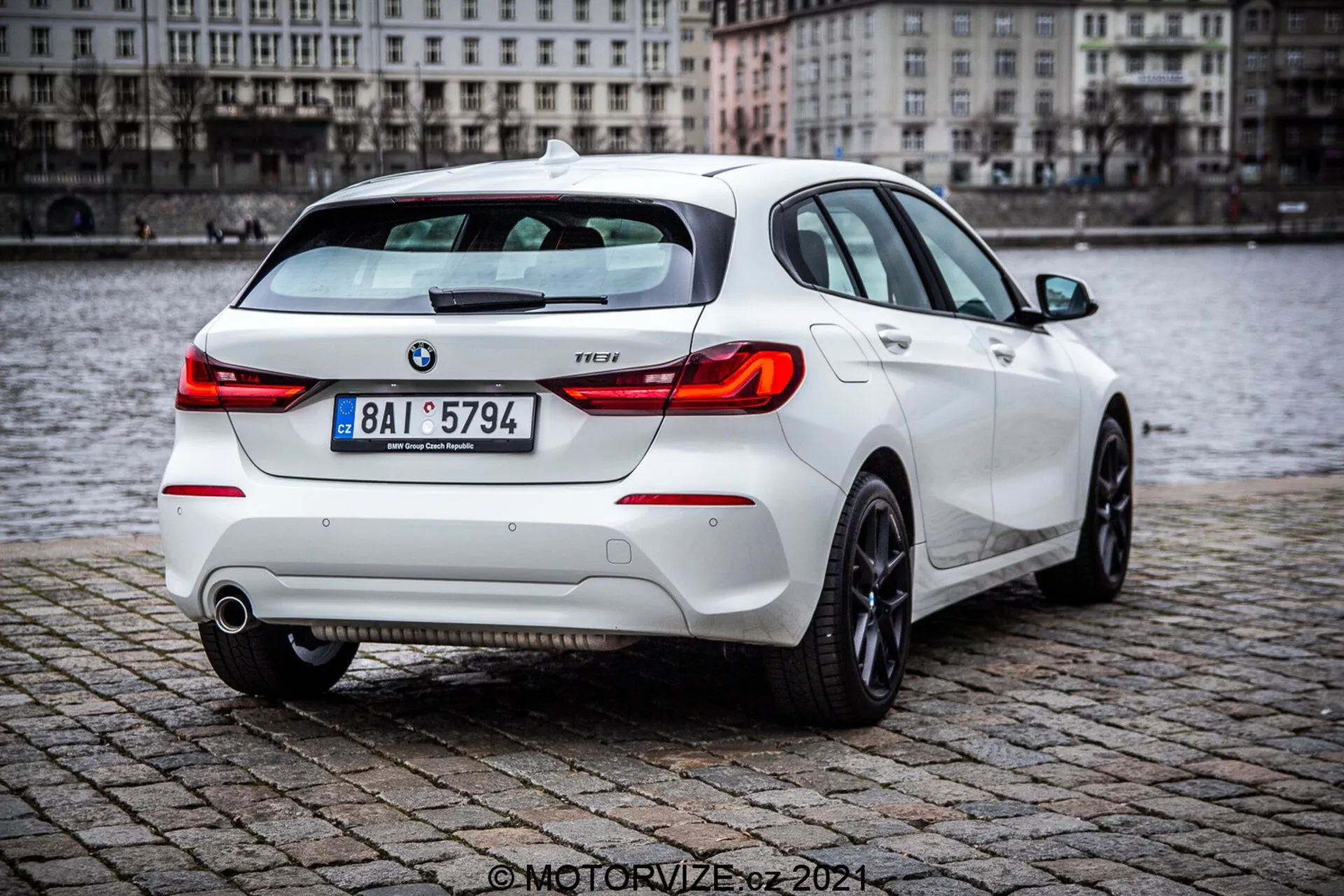 BMW serije 1 letnik 2019 (116i) bel hatchback, prikazan iz zadnjega tričetrtega kota s podrobnimi elementi vključujoč zadnje luči, pokrov prtljažnika, zadnji odbijač, izpušne cevi in del zadnjih koles, parkiran na mokri površini iz kock v bližini vodnega telesa. Slika izpostavlja športni zadnji spojler, moderne zadnje luči in vidne BMW in nivo opreme oznake.