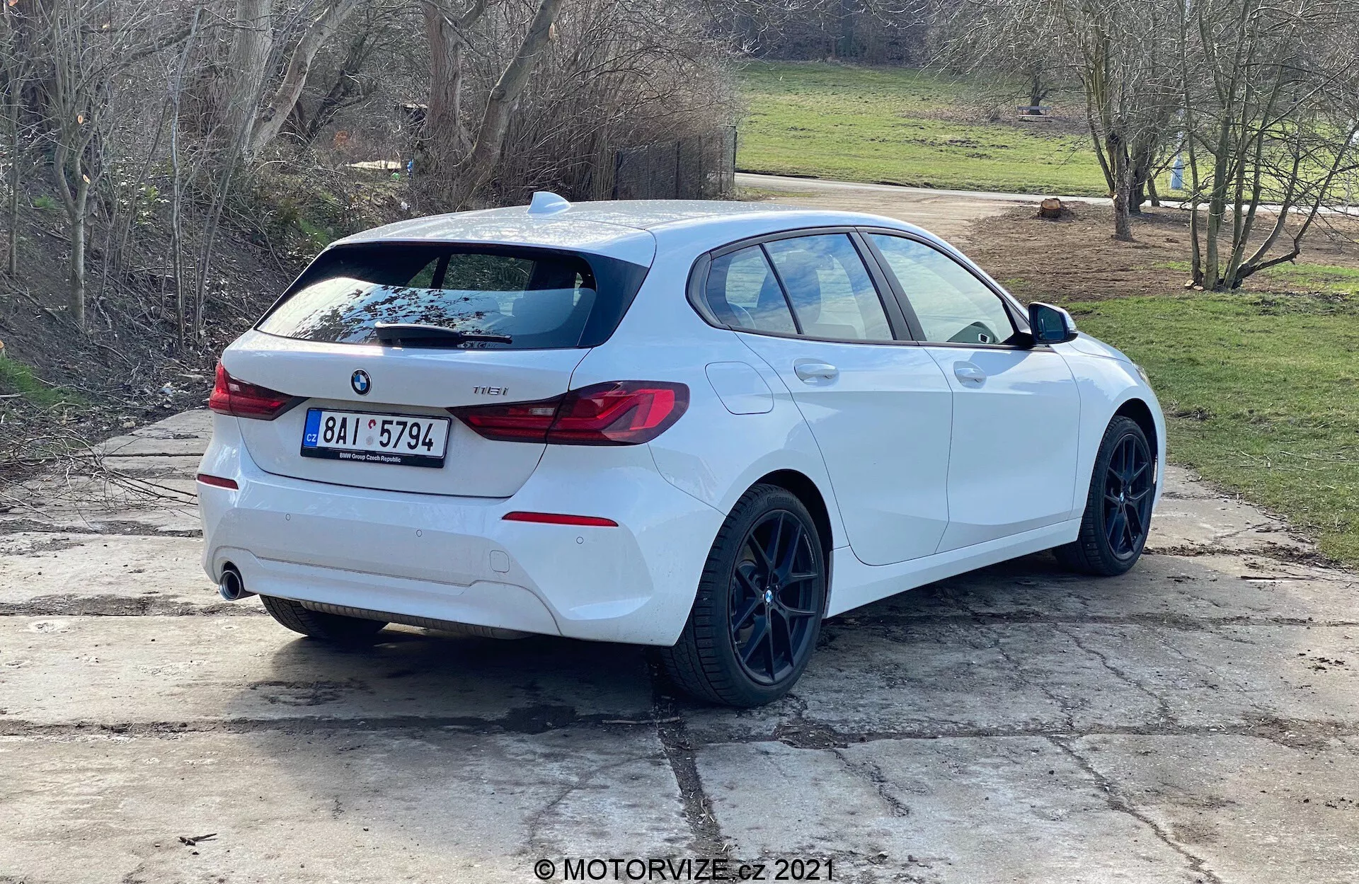 Zadnji trikotni pogled na bel BMW serije 1 (model BMW 116i 2019) parkiran na prostem v dnevni svetlobi. Bočni profil in zadek vozila sta jasno vidna, z emblemi BMW, oznako modela, značilnimi zadnjimi lučmi, zadnjim spojlerjem, črnimi platišči, hatchback obliko karoserije, dvojnimi izpušnimi cevmi, zadnjim brisalcem, rahlo zatemnjenimi okni in vzvratnimi ogledali z integriranimi smerniki. Vozilo je postavljeno na betonski površini s travnatimi deli in podeželskim ali predmestnim ozadjem.