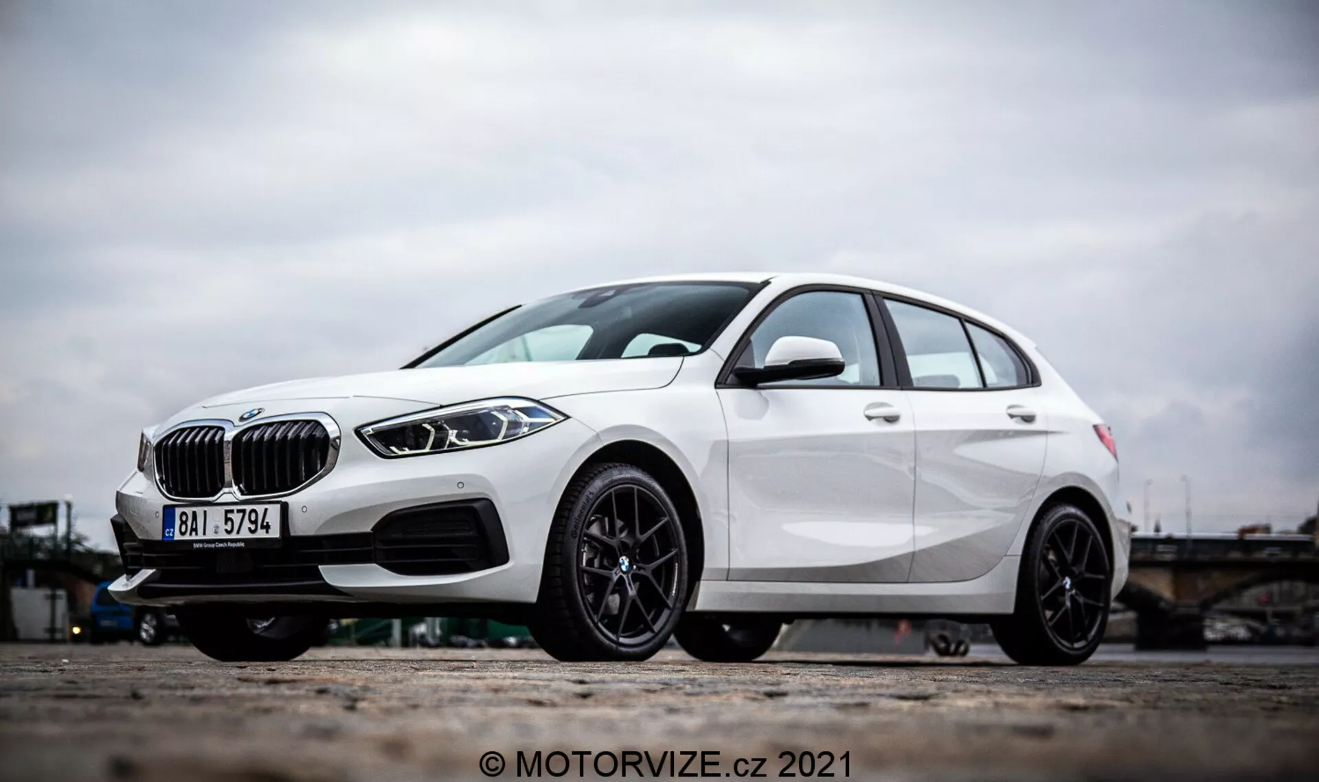 Poševni stranski pogled na bel BMW serije 1 2019 (BMW 116i), ki izpostavlja izrazite oblikovne elemente, kot so ledvičasti rešetki, elegantni LED žarometi in prednji odbijač z dovodi zraka in morebitnimi radarskimi senzorji za pomoč pri vožnji. Fotografija se osredotoča na lita platišča in spodnji del vozila ter poudarja športno zasnovo črnega večkraka in zmogljivo zavoro z diskom. Karakterne linije in aerodinamično oblikovanje hatchbacka še bolj poudarjajo njegov sodoben in dinamičen zunanji videz.