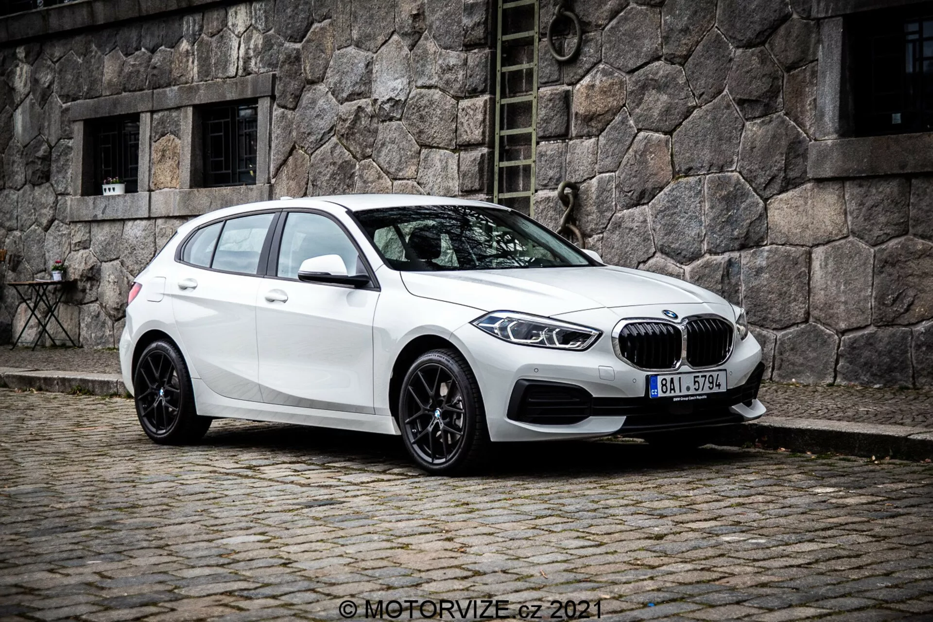 Bel BMW 1er-Reihe 2019 (BMW 116i) hatchback pogledan iz tretjinsko sprednjega kota, ki prikazuje levo stran vozila. Fotografija, posneta iz nekoliko nižje perspektive, poudari držo in oblikovne linije avtomobila. Postavljen je na tlakovani ulici z zgodovinsko kamnito zgradbo v ozadju, kar ustvarja klasično mestno sceno. Na sprednjem delu sta jasno vidna ledvična mreža in BMW emblem, obdana s prefinjenimi kotnimi LED žarometi. Avto ima večkraka črna aluminijasta platišča, padajočo strešno linijo, stranska ogledala v barvi karoserije z vgrajenimi smerniki ter zatemnjena okna za zadnje potnike.