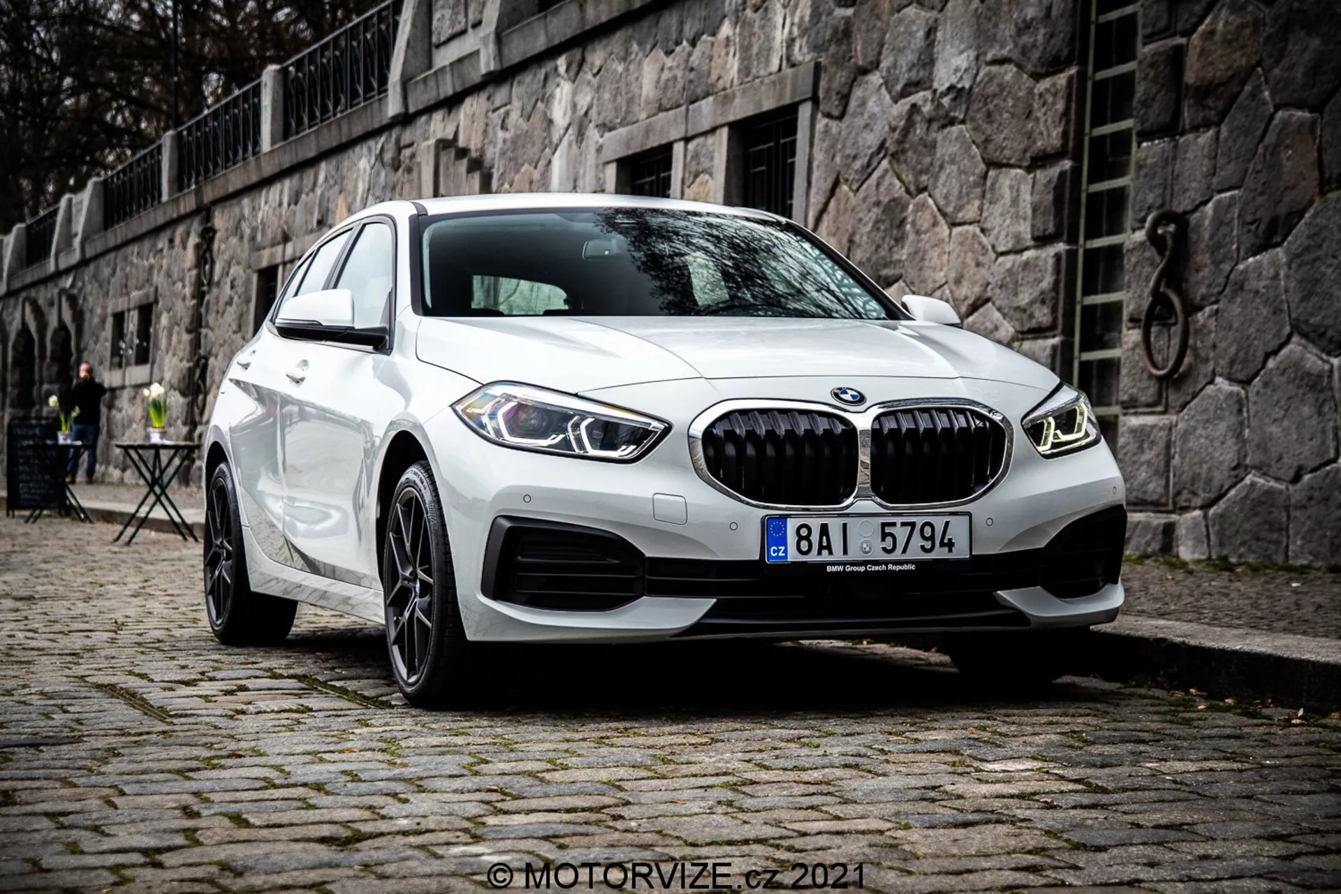 Sodobni beli BMW serije 1 (BMW 116i) model 2019, ki prikazuje značilno ledvično masko, dvojne okrogle dnevne LED luči in športno obliko sprednjega odbijača, parkiran na tlakovanem površju z zgodovinskim evropskim okoljem v ozadju, s kamnito steno in zunanjim pohištvom na kaviču.