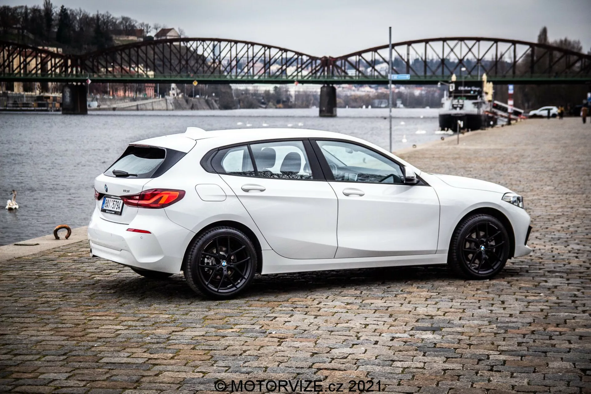Slika belega hatchbacka BMW 1er-Reihe 116i iz leta 2019 iz zadnje trikotne perspektive, poudarja njegovo elegantno, moderno obliko s športno držo, črnimi večkrakimi aluminijastimi platišči, rahlo zatemnjenimi okni in praktičnimi funkcijami, kot sta odmrzovalnik zadnjega okna in brisalnik. Avto prikazuje ostre linije in konture, dvojni izpušni sistem, subtilen zadnji strešni spojler, zadnjo kamero in parkirne senzorje. Parkiran je na kamnitem površju v bližini vodnega telesa z urbanim mostom v ozadju, kar nakazuje kontekst zunanjega življenjskega sloga.