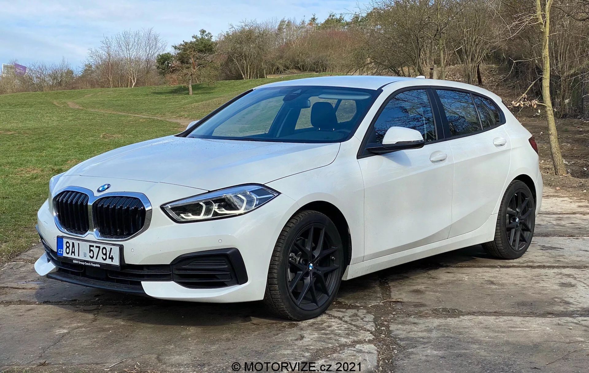 Slika prikazuje sodoben beli hatchback BMW serije 1 (BMW 116i), postavljen na prostem z naravnim, travnatim ozadjem. Posnetek iz treh četrt čelnega zornega kota, ki favorizira voznikovo stran, prikazuje značilno ledvičasto masko vozila, elegantne, kotne LED žaromete, črna lita kolesa, dobro definirano bočno gubo in vgrajene kazalnike smeri na stranskih ogledalih. Sprednji odbijač vključuje parkirne senzorje, medtem ko je mogoče sklepati na panoramsko stekleno streho in morebitno sprednjo kamero. Avto je parkiran na tlakovani površini in zaseda večji del okvira, kar nakazuje sodoben avtomobilski dizajn in napredne tehnološke funkcije.
