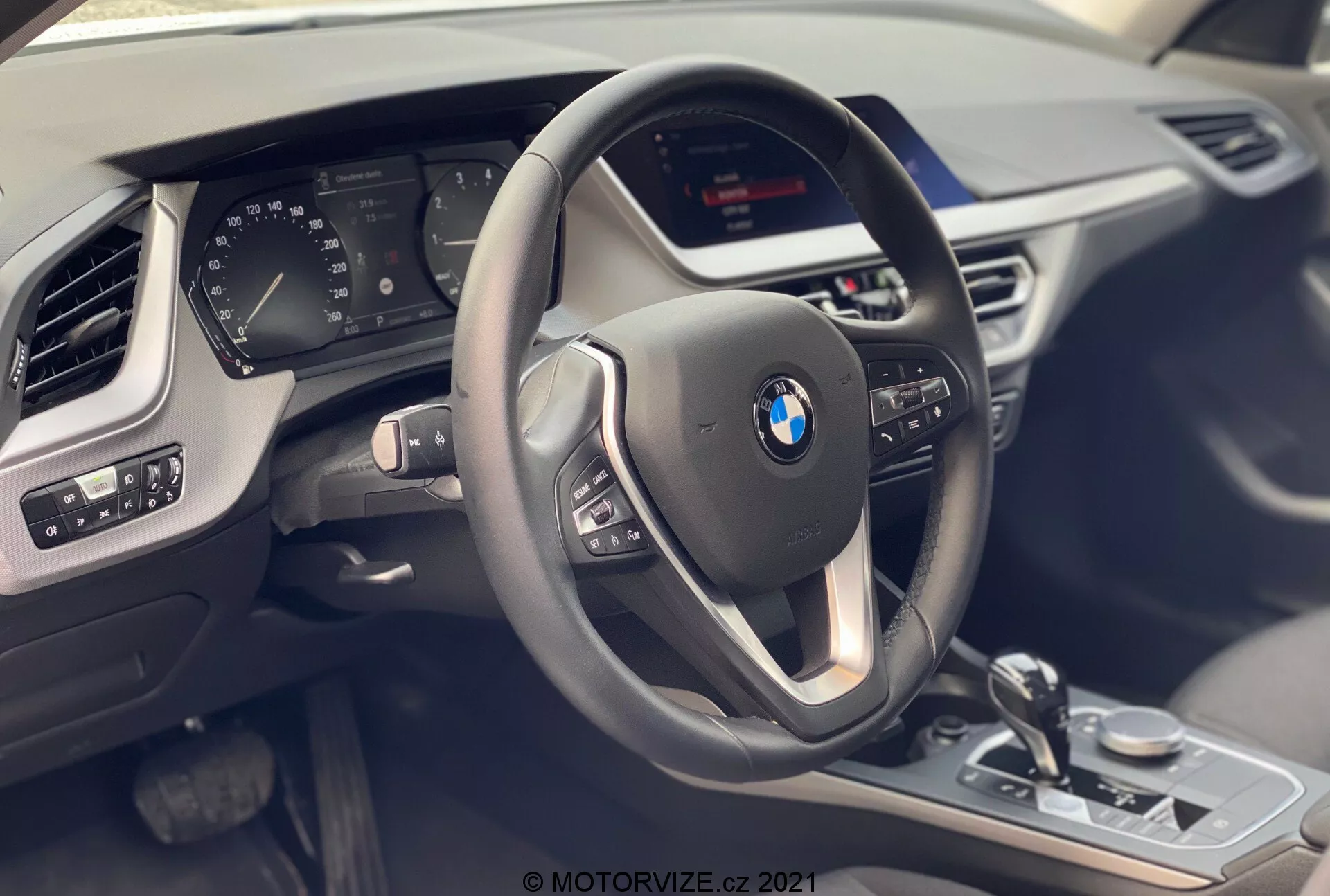 Notranji pogled na BMW serije 1 2019 (BMW 116i) s sedeža sovoznika, ki poudarja voznikov kokpit z modernim trikrako volanom z multifunkcijskimi gumbi in usnjeno prevleko s kontrastnimi šivi. Za volanom je delno viden digitalni instrumentni panel, ki prikazuje različne parametre vozila. Sredinska konzola vsebuje neviden zaslon za informacijsko-razvedrilne vsebine in vidne kontrole, kot so prestavna ročica, kontrolni gumb iDrive, izbirnik voznih načinov in kontrole parkirnih senzorjev, vse z metalnimi poudarki in sijočim črnim zaključkom. Plošča na voznikovih vratih prikazuje nastavitve ogledal in kontrole električnih oken. Vidna je tudi del zračne reže na sovoznikovi strani in tekstura armaturne plošče, kar poudarja luksuz, tehnologijo in sodobno oblikovanje.